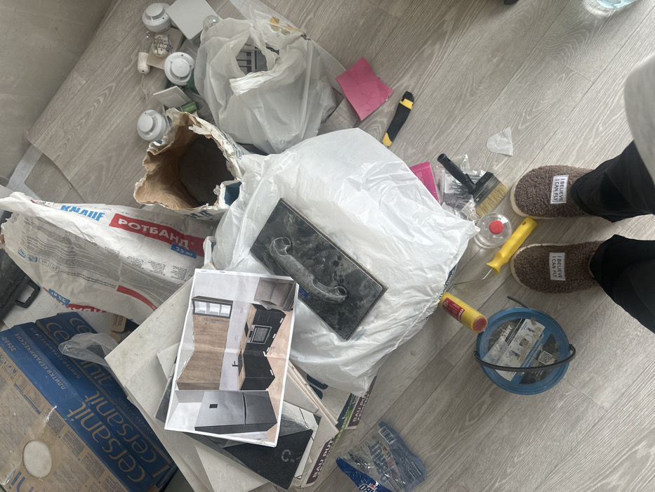 The scene depicts a ground-level view within a room in the city of Tankist, Russia, consistent with a renovation project. The floor, appearing to be light wood-effect laminate or vinyl, is extensively covered with construction materials and tools. On the left, several stacked cardboard boxes are visible, bearing "cersanit" branding and "плитки керамические" (ceramic tiles) text. Towards the center-right, an open paper bag labeled "KNAUF РОТБАНД" (a plaster mix) contains grey powdery material.
Central to the foreground is a printed architectural rendering of a modern kitchen design, featuring black lower cabinetry, wooden upper cabinets, a black refrigerator, a sink, and an oven, resting on a white plastic bag. A dark grey trowel is positioned atop this bag. Surrounding these items are scattered objects including several white circular components (possibly smoke detectors or recessed light fixtures), a yellow-handled utility knife, a paint roller, a paintbrush, a blue spool-like container, and a pink piece of paper.
In the immediate foreground, the lower legs and feet of an individual are visible, wearing brown fluffy slippers. Each slipper has a white rectangular tag with the text "I BELIEVE I CAN FLY" embroidered in black. No direct interactions are depicted beyond the implied presence of the individual in the cluttered environment.