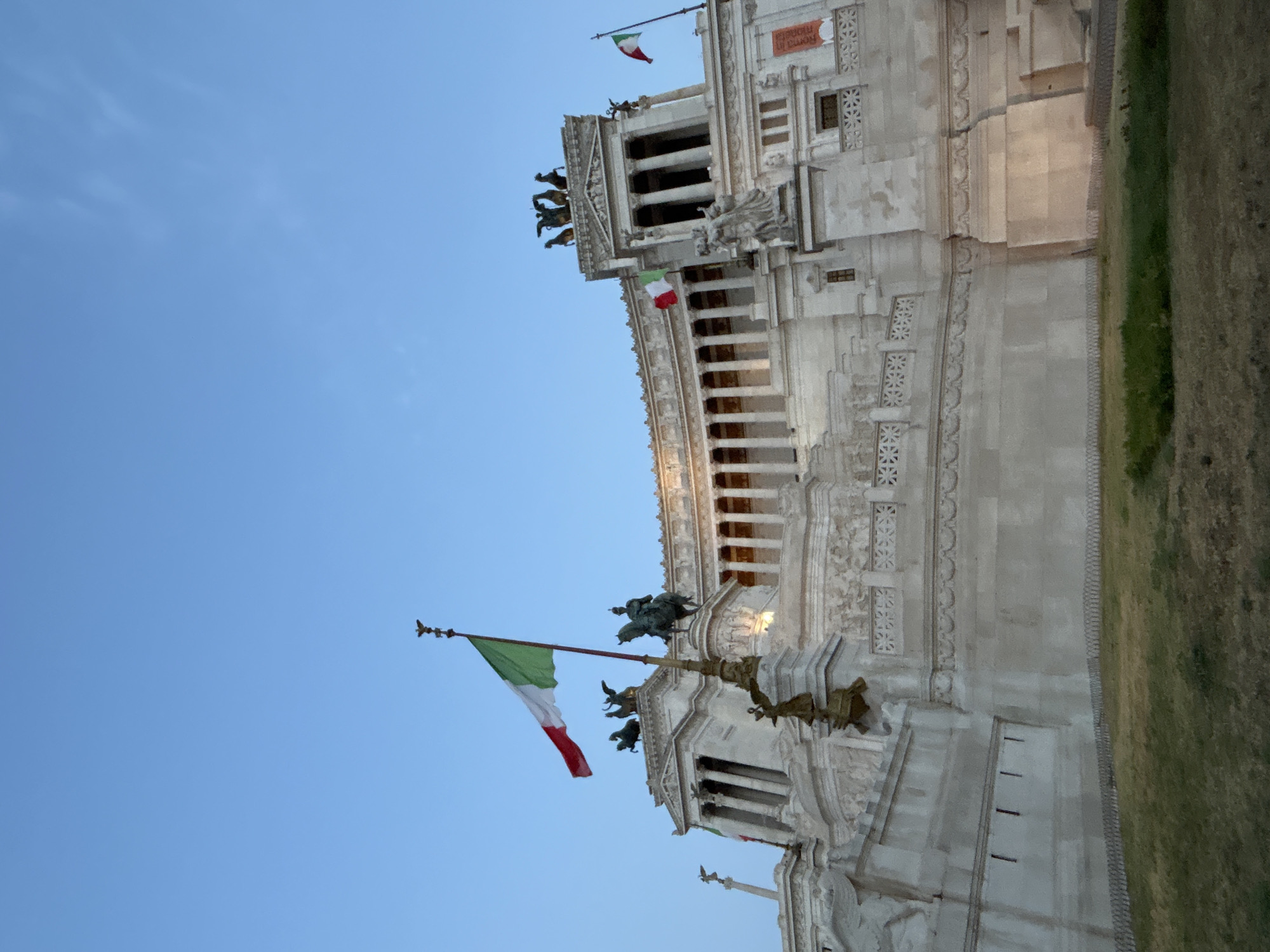 The image captures a low-angle, exterior view of the Altare della Patria, also known as the Victor Emmanuel II National Monument, in Rome, Italy, against a clear blue sky. The monument, constructed of white marble, features grand architectural elements such as columns, intricate carvings, and statuary. Prominently displayed are several Italian flags, with one large flag waving from a flagpole in the foreground, and smaller flags positioned on various parts of the building. Statues of horses and figures adorn the upper levels of the monument. In the foreground, a grassy area with some dry patches slopes upwards towards the base of the monument, separated by a low metal fence. The lighting suggests it is either early morning or late afternoon. There are no visible people in the image.