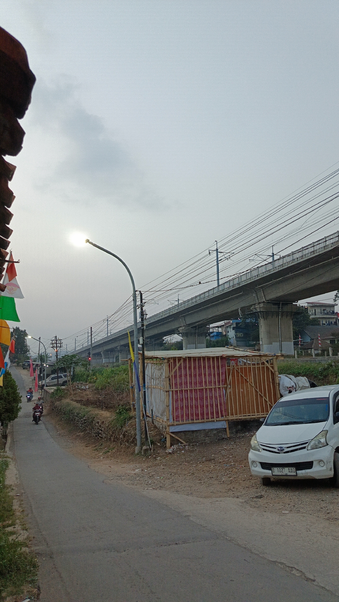 The image depicts a scene in Margajaya, Indonesia, featuring a road winding upwards towards a massive overpass. The sky is overcast, with the sun partially visible, creating a hazy light.
On the left side of the road, there are colorful flags, possibly decorations, hanging from what appears to be a building's roofline. Further up the road, three motorcycles are in motion, with riders visible. A few trees and some greenery line the road.
Dominating the upper portion of the image is a long, concrete overpass, likely for a railway or highway, supported by numerous concrete pillars. Overhead power lines stretch across the sky, following the path of the overpass.
On the right side of the road, a small, makeshift structure with bamboo framing and walls covered in a red and white patterned material is situated. Next to this structure, a white minivan is parked. The ground is a mix of dirt, gravel, and some debris. The overall environment suggests a semi-urban or developing area.