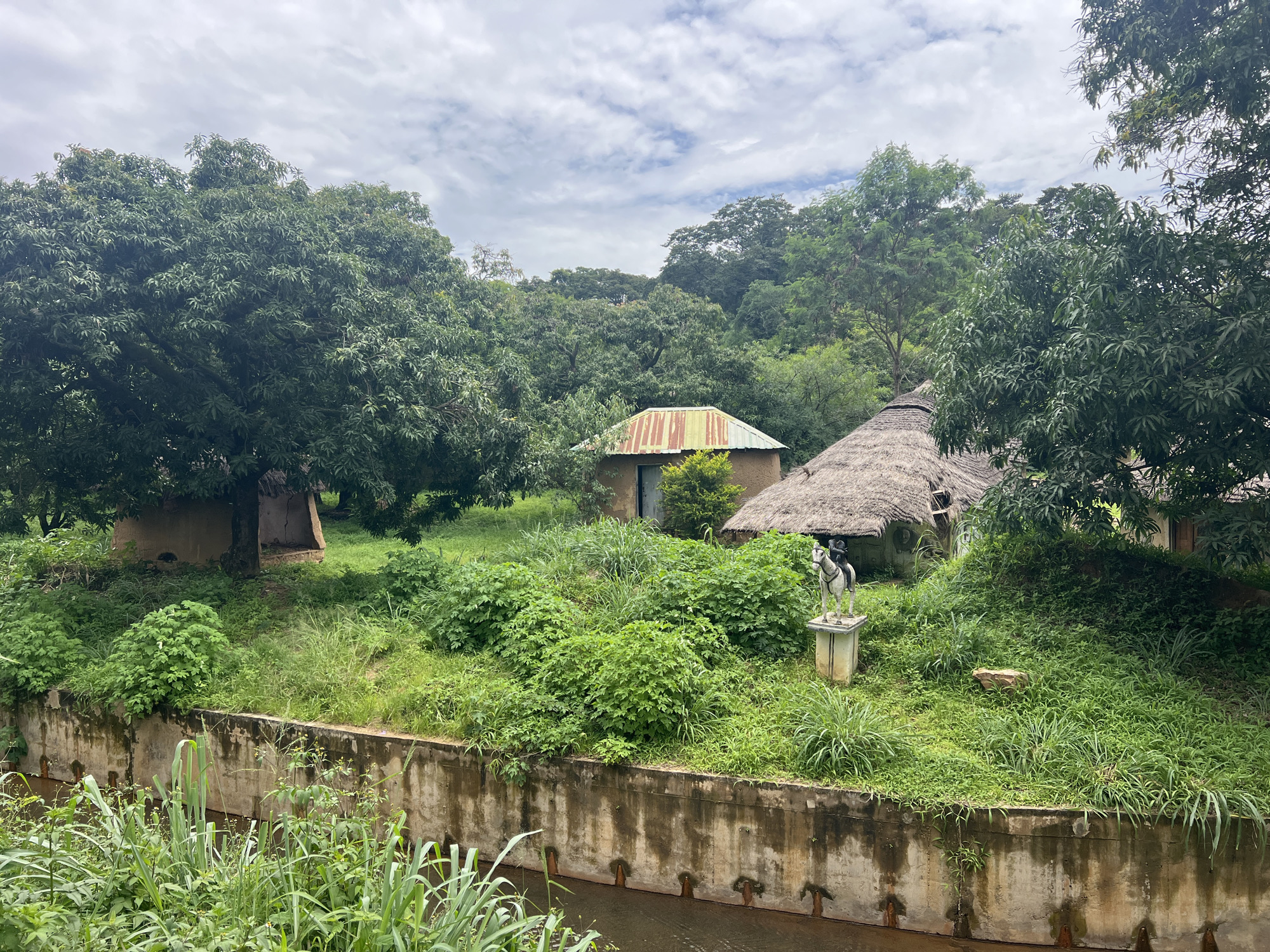 The image depicts a scene within the habitat area of the zoo in Jos, Nigeria. Two rustic structures are visible, one with a corrugated iron roof and the other with a thatched roof, suggesting an African village-style enclosure. In front of these structures, a statue of a person on horseback stands on a pedestal amidst lush green vegetation. The background is dominated by dense trees, creating a naturalistic environment. A concrete wall borders a body of water in the foreground, with tall grasses growing along its edge. The sky above is partly cloudy. The scene appears to be a static display within the zoo, designed to showcase a particular cultural or environmental theme.