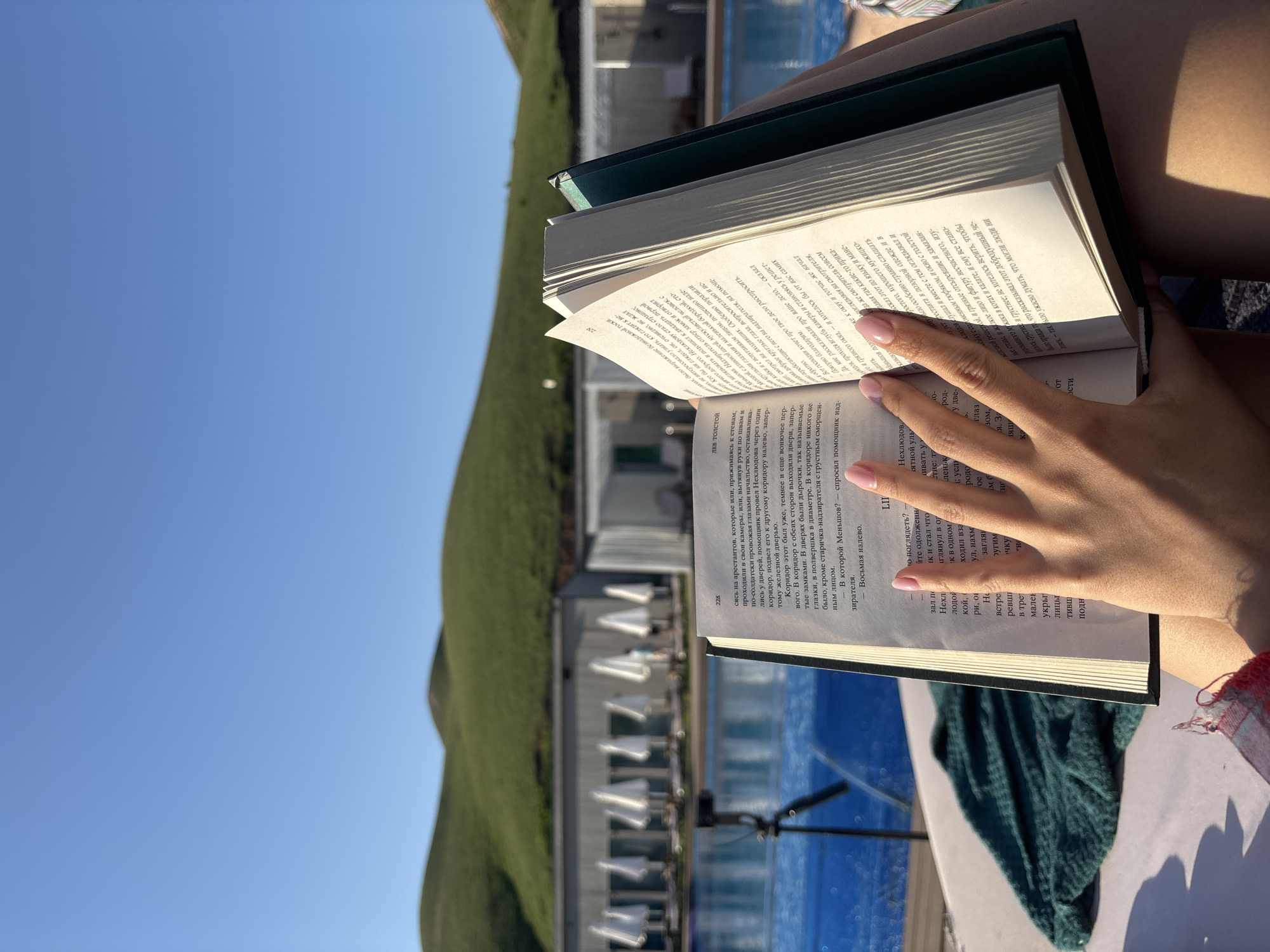 The image captures a close-up of a person's hands holding an open book, likely while relaxing outdoors by a pool. The scene is set under a clear, bright blue sky, suggesting sunny daytime. In the midground, a vibrant blue swimming pool glistens in the sun, with a row of resort-style buildings featuring white awnings visible behind it. Lush, green hills rise in the background, adding to the scenic beauty of the location, which is provided as Дрокино, Russia.
The primary activity is reading, indicated by the person's hands holding the book. The hands have light pink nail polish. Strong sunlight casts shadows on the book's pages and the person's hand. The book has a dark green cover and is open to pages filled with Russian text. Visible text includes page numbers "228" and "229", and the OCR clearly identifies "ЛЕВ ТОЛСТОЙ" (Leo Tolstoy) as the author. A turquoise towel or piece of fabric is partially visible beneath the book. The overall impression is one of leisure and tranquility, enjoying a book in a pleasant, sunny environment.