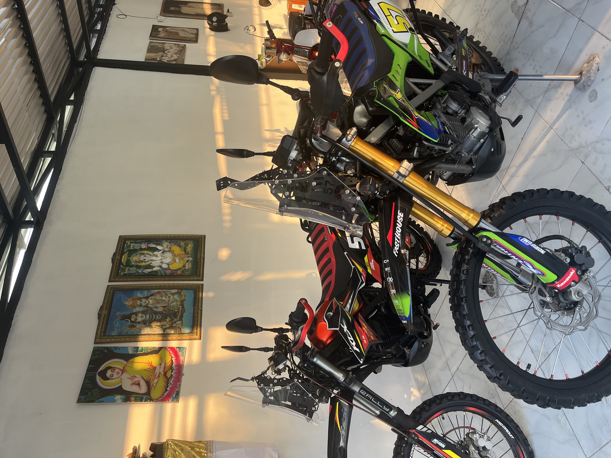 The image captures an indoor space, likely a garage or private dwelling in Tegallalang, Indonesia, illuminated by warm, directional sunlight. The setting features multiple parked motorcycles against a white wall, suggesting either early morning or late afternoon due to the long shadows and golden light.
In the foreground, two off-road motorcycles dominate the scene. One is a green, black, and blue Kawasaki dirt bike, prominently displaying text such as "Kawasaki," "FASTHOUSE," "NORIFUMI," and the racing number "5." Its front wheel, disc brake, and gold-colored suspension forks are visible. Adjacent to it is a red, black, and yellow bike, which has "RALLY" printed on its suspension and "PRO CIRCUIT" on its wheel rim, along with the number "57." In the background, a classic red scooter or motorcycle is partially visible.
The white wall behind the motorcycles is adorned with three framed religious pictures. The topmost depicts the Hindu deity Ganesha, the middle frame shows Shiva and Parvati with Ganesha, and the bottom frame features a meditating Buddha. These artworks are bathed in the golden light.
The architecture includes a visible ceiling structure with dark, exposed metal beams and slatted roofing on the left side of the frame. The floor is covered with light-colored tiles. Additional details include a black motorcycle helmet resting on the floor in the background and a small table draped with gold fabric in the bottom left. Other visible text includes "MOTOCROS" and "ENTHAL." No specific activity is depicted, as the bikes are stationary.
