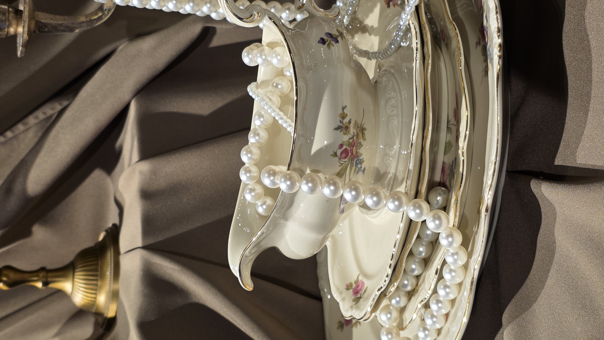 The image depicts a still life arrangement of antique porcelain tableware, pearl necklaces, and a brass candlestick against a backdrop of draped brown fabric. The porcelain items, including a gravy boat and plates, are decorated with floral motifs and gold trim. The pearl necklaces are draped over the tableware and extend towards the candlestick. The brass candlestick, partially visible on the right side of the frame, is ornate and appears to be an antique piece. The fabric background is softly folded, creating shadows and highlighting the textures of the objects. The lighting is warm, suggesting an indoor setting with natural or artificial light casting gentle shadows. There are no people, actions, or interactions depicted. The location is not identifiable within the city of Riga, Latvia, or any other specific geographical area.