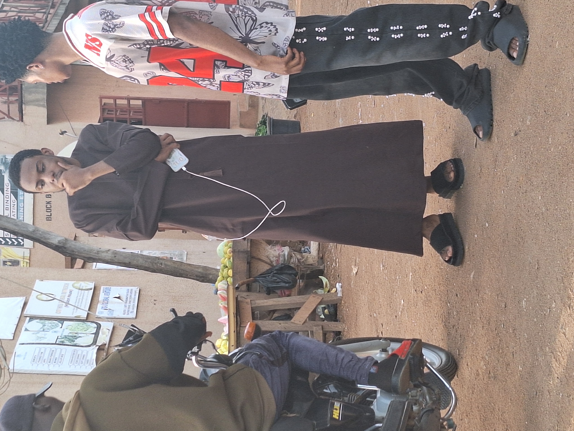 Here is a description of the image:
Location: Sabon Tasha, Nigeria.
A medium shot captures an outdoor scene with several individuals and objects. Two men are standing, the first clad in a white shirt with red accents and black pants with white embellishments. The second man wears a long brown robe and black sandals. A white cable extends from his pocket, connected to a device he holds.
In the foreground, a person is seated on a motorcycle, partly obscured, wearing a green top and jeans. To the side, a fruit stand displays various fruits. Adjacent to it is a wooden structure with posters and signs affixed. The ground is dirt or unpaved road. The background shows a building with a red door. Posters on the wall include the text "GLOBAL VENTURE" and "BLOCK B." The brand name HLXE is visible on the motorcycle.