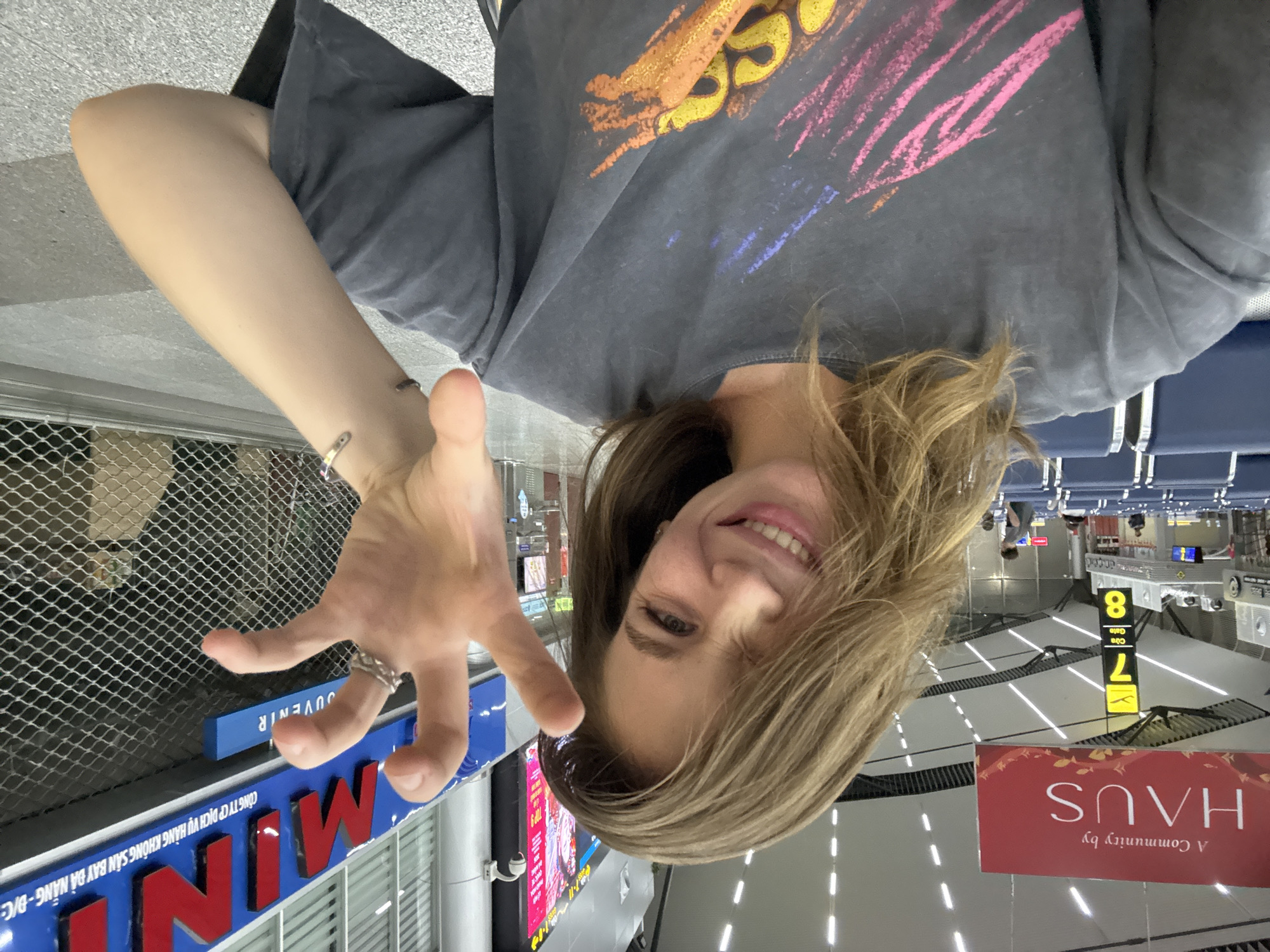 A young woman is taking a selfie in what appears to be an airport terminal. She has light brown hair and is smiling with one eye winking. She is wearing a dark grey t-shirt with a graphic print and has her right hand raised in front of her face, fingers splayed. In the background, there are rows of blue seats, a red sign that reads "A Community by HAUS", and a digital display showing gate numbers 7 and 8. Further in the background, a large blue sign with red lettering is visible, which appears to be a store name or advertisement, with Vietnamese text underneath. The overall environment suggests an indoor public space with commercial elements, consistent with an airport in Đà Nẵng, Vietnam.