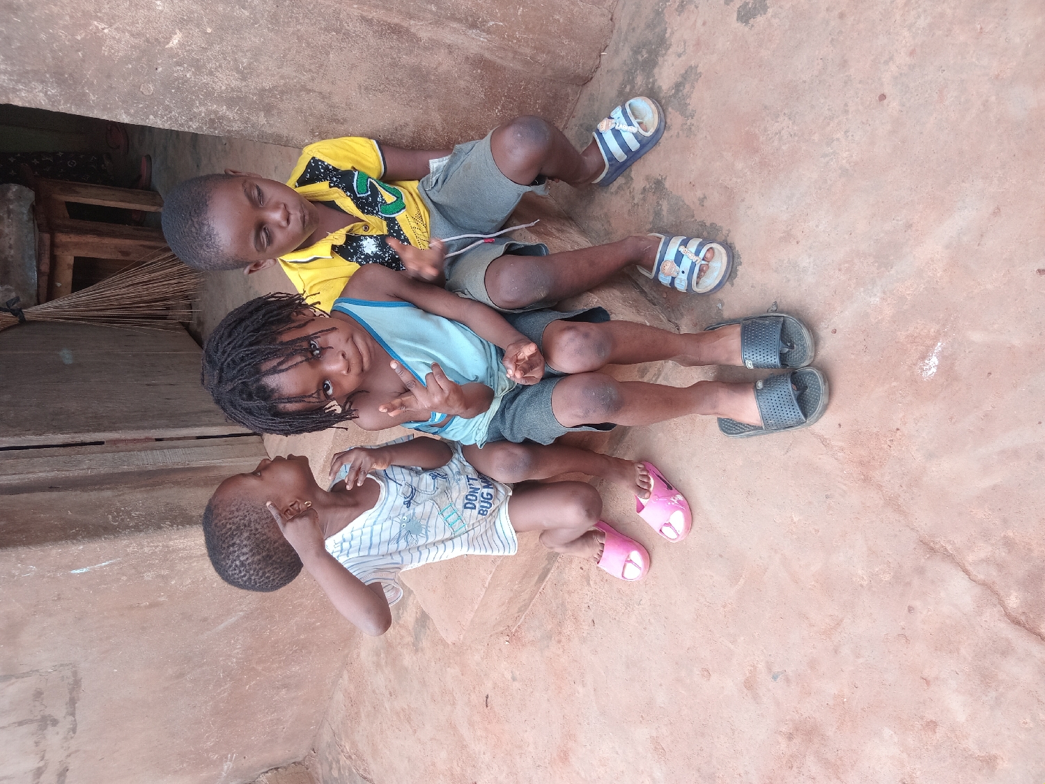 Three young children are depicted lying on a rough, earthy-toned ground or floor, likely outdoors or in a semi-enclosed humble setting in Shagamu, Nigeria.
The topmost child, appearing to be a boy, wears a yellow short-sleeved t-shirt with black designs and grey shorts. He has short dark hair and is wearing blue and white striped slide sandals. He lies on his back, looking directly towards the camera with a relaxed expression.
The middle child, also a boy with dark braided hair (dreadlocks), wears a light blue sleeveless tank top and grey shorts. He has dark grey/black slide sandals on his feet. He is also lying down, looking towards the camera and holding up two fingers in a peace sign.
The bottom child, the smallest of the group, has very short, dark hair and wears a white t-shirt with light blue stripes and some graphic elements. Part of a text "DON'T BUG M" (likely "DON'T BUG ME") is visible on his shirt. He is wearing pink slip-on sandals. He is lying on his side, looking towards the middle child, with one hand near his ear.
In the background, behind the children's heads, a wooden structure and a broom or bundle of sticks are visible. The ground appears to be made of concrete or dried mud, suggesting an informal dwelling or outdoor area. The lighting indicates it is daytime. The children appear calm and playful, engaging with the camera or each other.