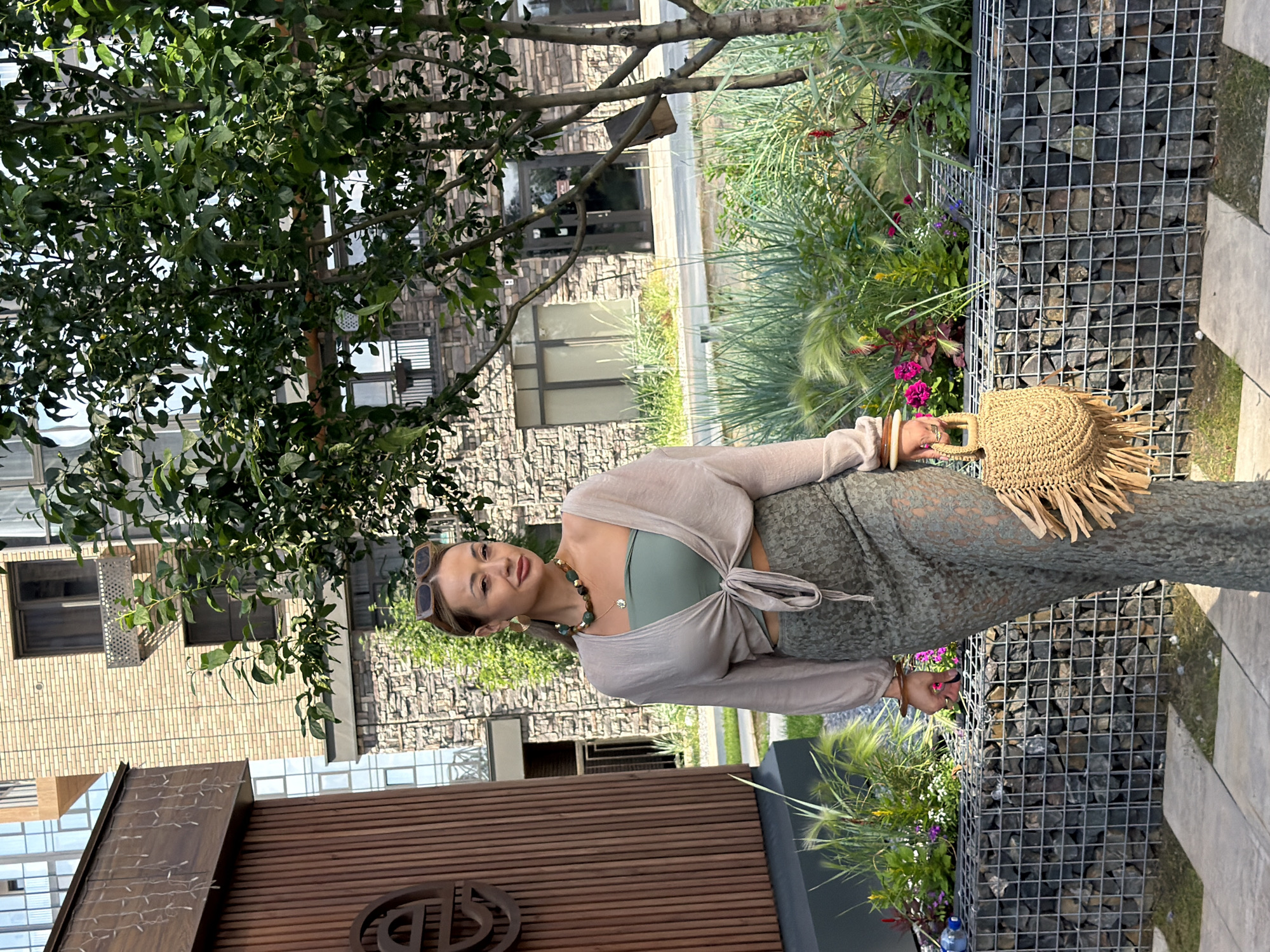 A woman stands in front of a modern building with a stone facade and wooden accents. She is wearing a green bandeau top, a beige open-front cardigan tied at the waist, and a long, green, lace-like skirt. She has on a statement necklace with large, multi-colored beads and gold hoop earrings. She is also wearing sunglasses on her head and carrying a straw handbag with fringe details. The background features a lush green tree and garden area with a gabion wall filled with stones. The scene appears to be outdoors in a contemporary urban setting, possibly a courtyard or plaza. There are no obvious indications of the specific location being Krasnoyarsk, Russia; the description is based solely on visual cues within the image.