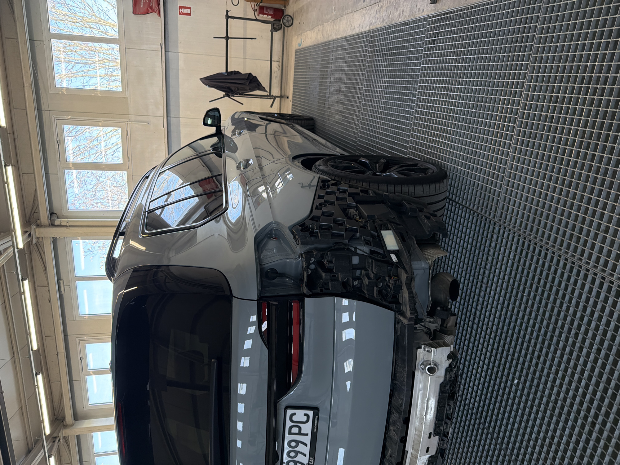 An overhead shot, rotated 90 degrees counter-clockwise, depicts a modern grey car undergoing repair in what appears to be an automotive workshop or garage. The vehicle, possibly an SUV or crossover, has its rear bumper removed, exposing the internal structure, wiring, and a single exhaust pipe on the right side. A black multi-spoke wheel with a tire is visible on the right rear of the car. The car's body is a solid grey color, and reflections of the ceiling lights and exterior windows are visible on its surface.
The setting features a distinctive dark grey metal grate flooring, typical of a service bay, stretching across the floor. White walls surround the space, with multiple rectangular windows higher up, revealing a clear daytime sky and bare tree branches outside. Overhead, long fluorescent light fixtures illuminate the area. In the background, against a white wall, a red fire extinguisher is mounted, with partially visible text "ПОЖАРОГАСИТЕЛ" (Bulgarian for fire extinguisher) and "10". A black stand with a cover, resembling an umbrella or equipment stand, is also present near the wall.
The car's license plate is clearly visible on the rear, displaying the characters "999 PC". Below the numbers, the text "www.bgcar.bg" is visible, along with the partially obscured word "CAR". The overall scene suggests a professional environment focused on vehicle maintenance or bodywork.