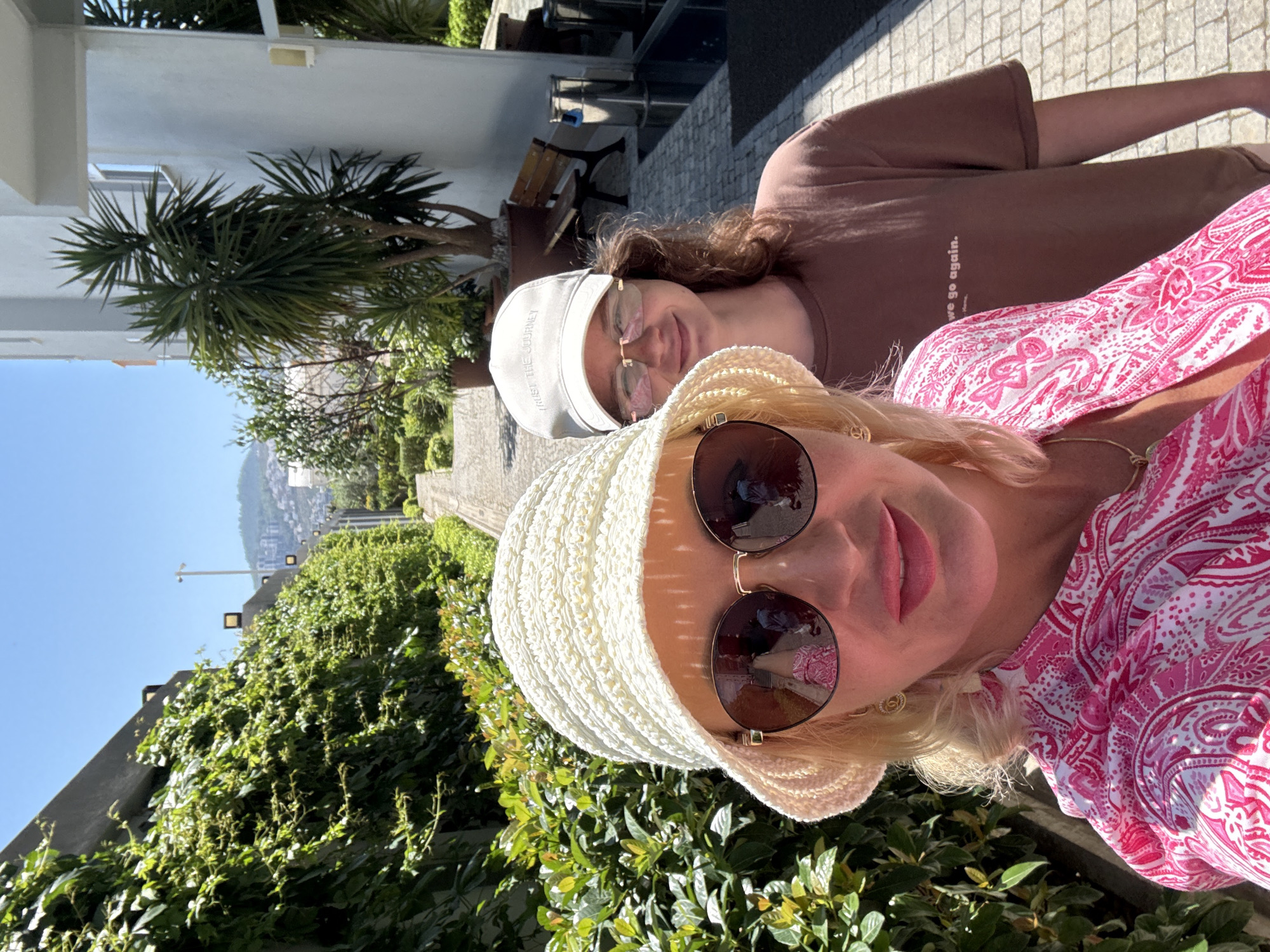 Two women are posing for a selfie outdoors. The woman in the foreground has short blonde hair, large sunglasses, and a white crocheted hat. She is wearing a pink paisley-patterned top. Behind her is a younger woman with brown hair, glasses, and a white baseball cap. She is wearing a brown T-shirt. The background shows greenery and a cityscape with buildings visible on a hill. The location is Unknown, Unknown.