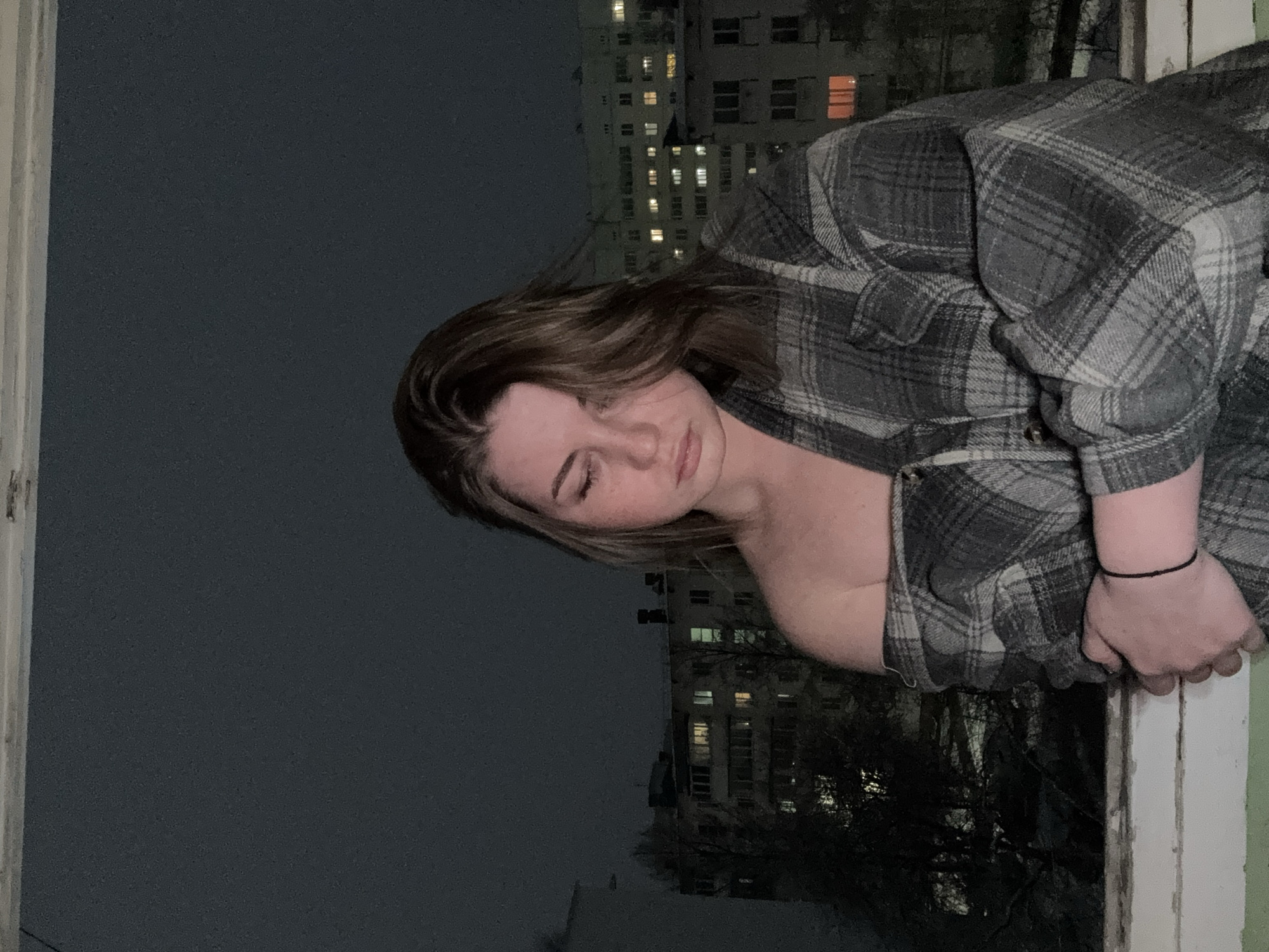 A young woman leans out of a window at night. She is wearing a grey and black plaid shirt. The city skyline is visible in the background, with lights on in some buildings. The location is Chaikovsky, Russia.