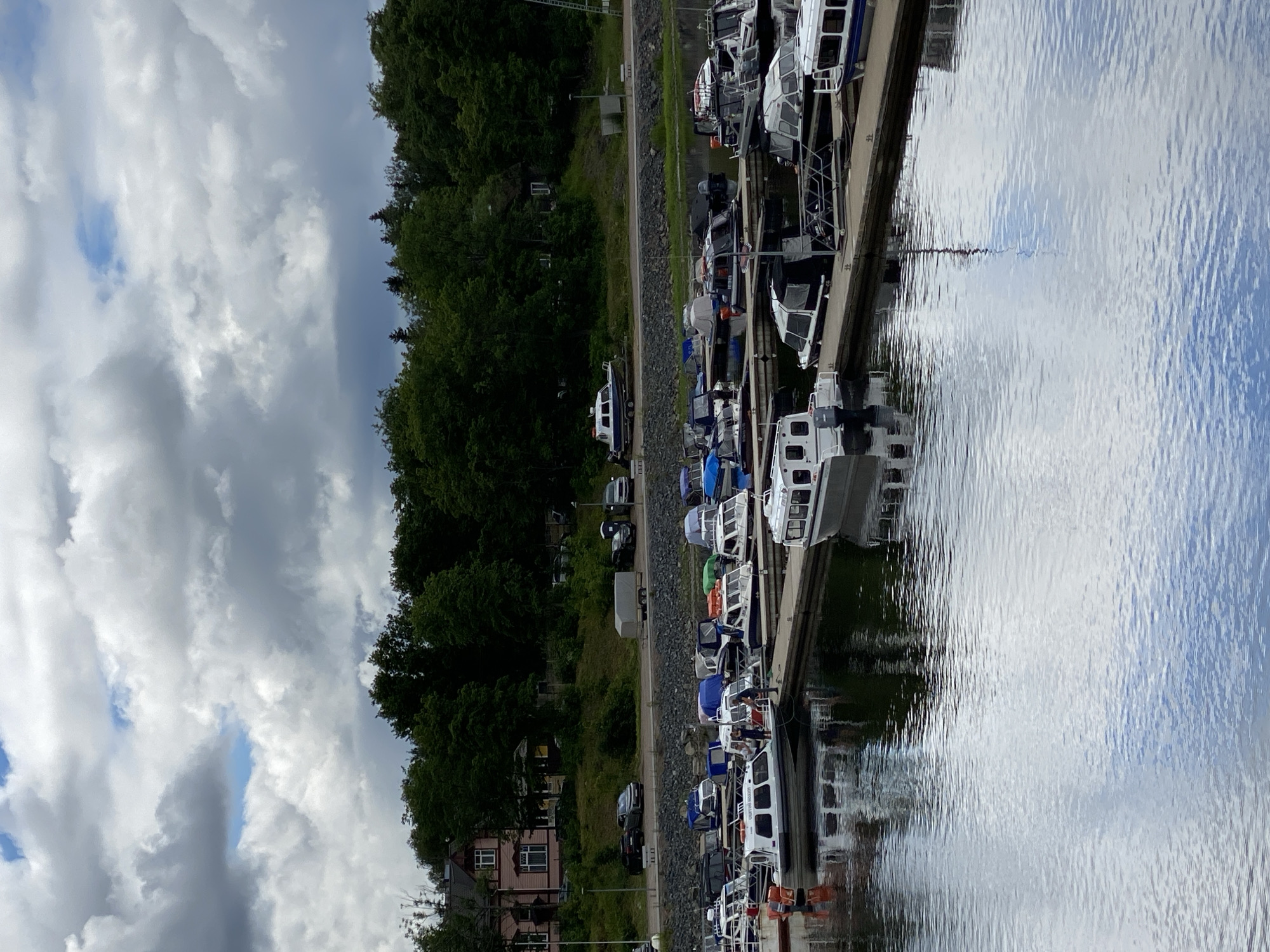 The media file shows a row of boats docked along a pier on a body of water. The boats are varied in size and color, with many appearing to be modern motorboats. Some have enclosed cabins, while others are open. Several boats are white, and some have blue or orange accents. A few people are visible on the decks of the boats, suggesting they are either arriving, departing, or simply enjoying a day out.
The setting is a marina, with a tree-lined shore visible behind the pier. The trees are lush and green, indicating it is likely summer or late spring. The water is calm and reflects the sky, which is partly cloudy with patches of blue visible. The overall atmosphere appears peaceful and relaxed. A small building, possibly a house or part of the marina facilities, is visible near the shore.
The time of day is difficult to ascertain precisely due to the cloud cover, but the lighting suggests it is daytime, perhaps late morning or afternoon. There are no overt signs of specific events, but the presence of multiple boats and people suggests a leisurely activity, such as boating or fishing. No significant text is visible in the image. The location context provided is Sortavala, Russia, which aligns with a coastal or lakeside setting.