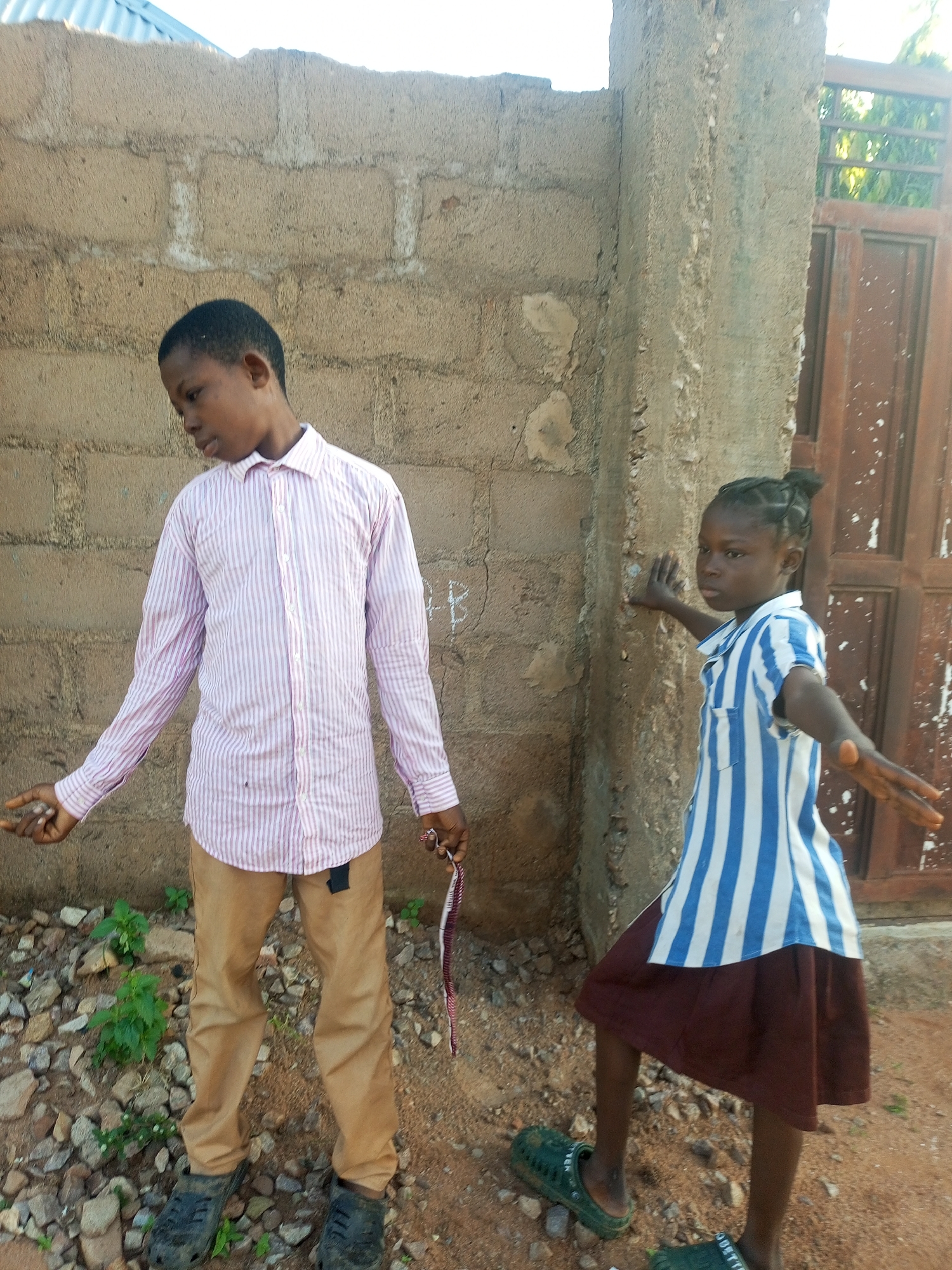 The image shows two young children standing outdoors in what appears to be Tempure, Nigeria. A boy on the left, wearing a pink and white striped long-sleeved shirt and tan pants, stands facing left with his head tilted down. He is holding a thin, red and white object in his right hand. To the right of the boy, a girl in a blue and white striped short-sleeved shirt and a dark red skirt stands facing forward, her arms outstretched. Both children are barefoot, with the boy wearing dark clogs on his feet and the girl wearing green clogs. They are standing on a surface of gravel and dirt, with a textured, light brown brick wall behind them. To the far right, a portion of a reddish-brown metal gate with vertical bars is visible, set against a concrete pillar. The lighting suggests it is daytime.
