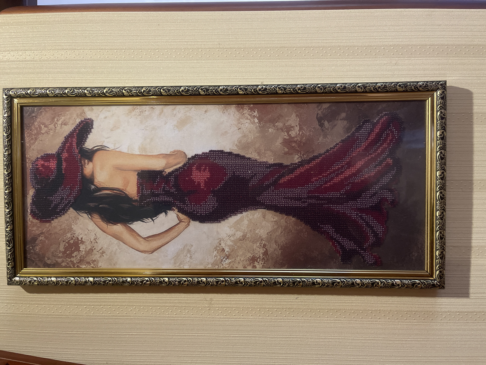 A framed diamond painting depicts a woman from the back, wearing a large, wide-brimmed red hat and a floor-length red gown. The painting is hanging on a light beige wall with a subtle vertical textured pattern. The frame is ornate, with a gold finish and intricate dark floral detailing. The woman's long, dark hair cascades down her back, partially obscured by the hat. Her skin tone is fair, and she is shown with one hand on her hip. The gown is fitted at the bodice and flares out dramatically towards the hem, featuring a diamond pattern that creates a shimmering effect. The background of the painting is abstract, with muted brown and beige tones, giving a sense of depth and texture. The lighting casts a soft shadow on the wall to the right of the painting.