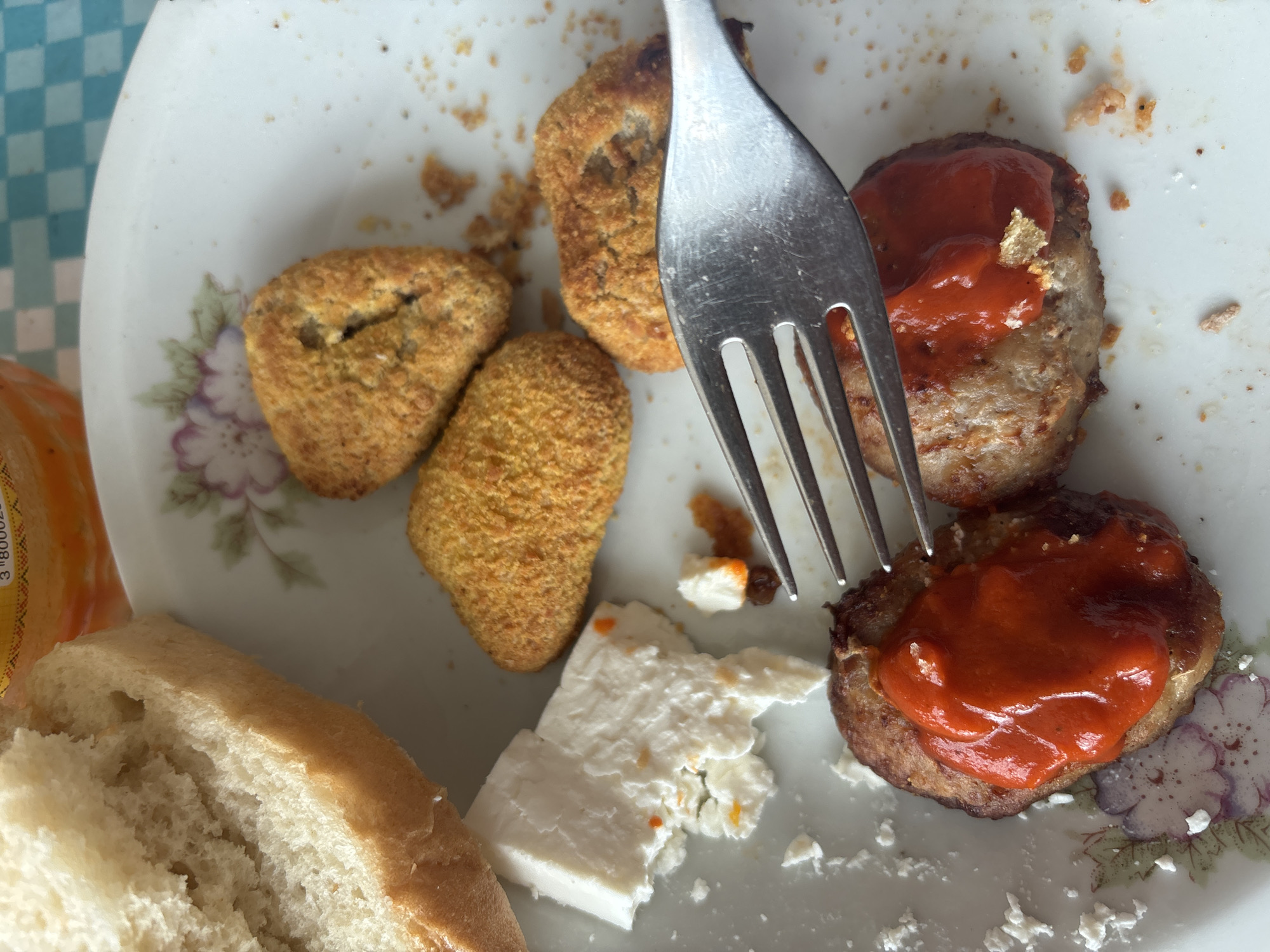 A cluttered plate on a table in an unspecified location, likely Zlatograd, Bulgaria, contains remnants of a meal. The plate is white with floral decorations. The meal appears to include:
* Several breaded, fried items (possibly chicken nuggets or similar).
* Two patties topped with red sauce (possibly ketchup or a tomato-based sauce).
* A chunk of white cheese.
* A piece of bread.
* A metal fork is present, partially obscured by the food.
* The background suggests a light-colored tablecloth, potentially checkered.