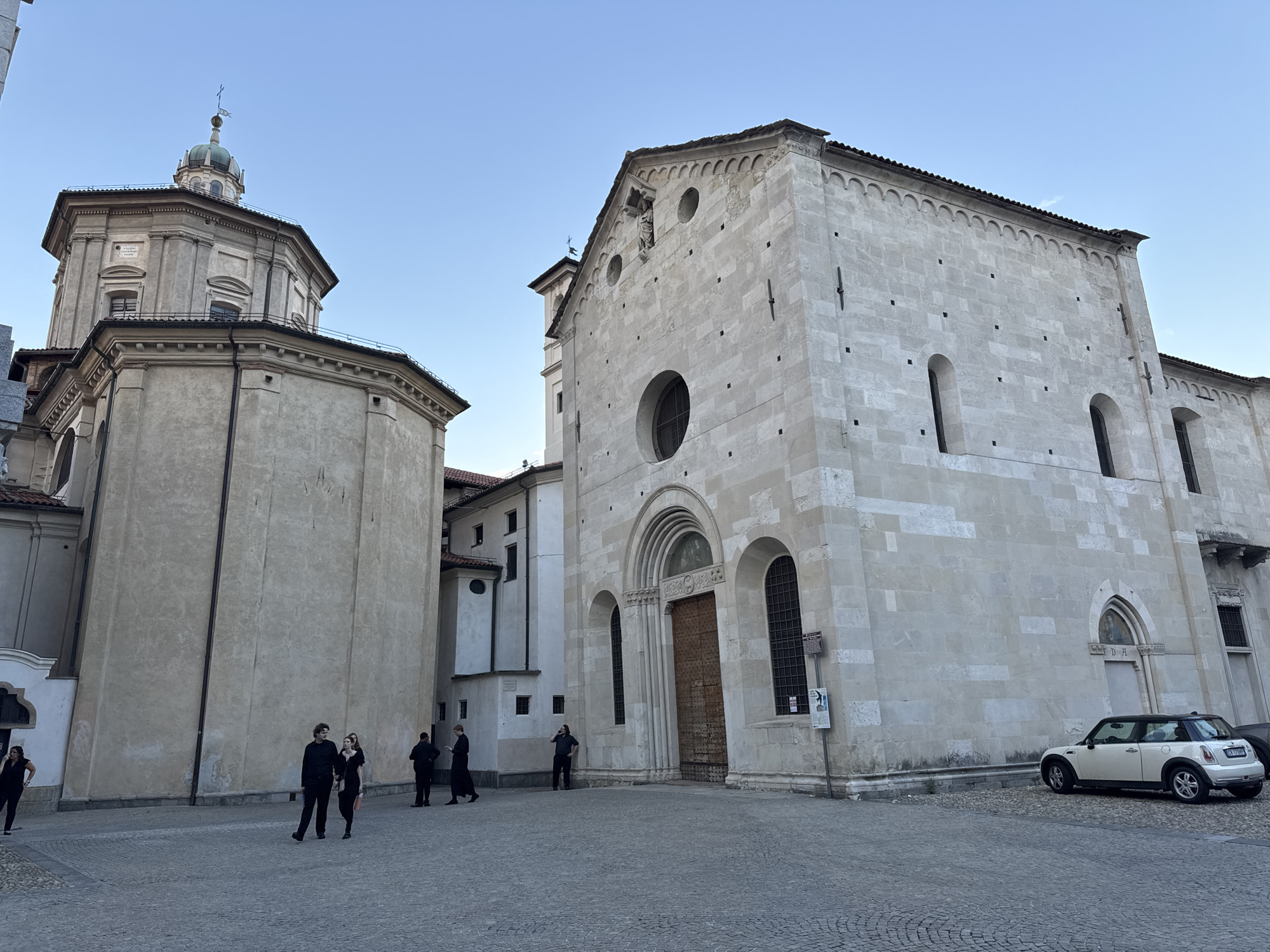 In Varese, Italy, on a cobblestone plaza, a cream-colored Mini Cooper is parked next to a large, light-stone building, possibly a church or cathedral. To the left of this building is a taller, older structure with a domed roof topped with a small cross. The larger building features a large, arched wooden door and several tall, arched windows. Above the door, a circular window is visible, and a small statue is set into the pediment. The facade is constructed of light-colored, rectangular stone blocks. Several individuals in dark clothing are scattered across the plaza, with two couples walking and one person standing near the building's entrance. The sky is clear and blue.