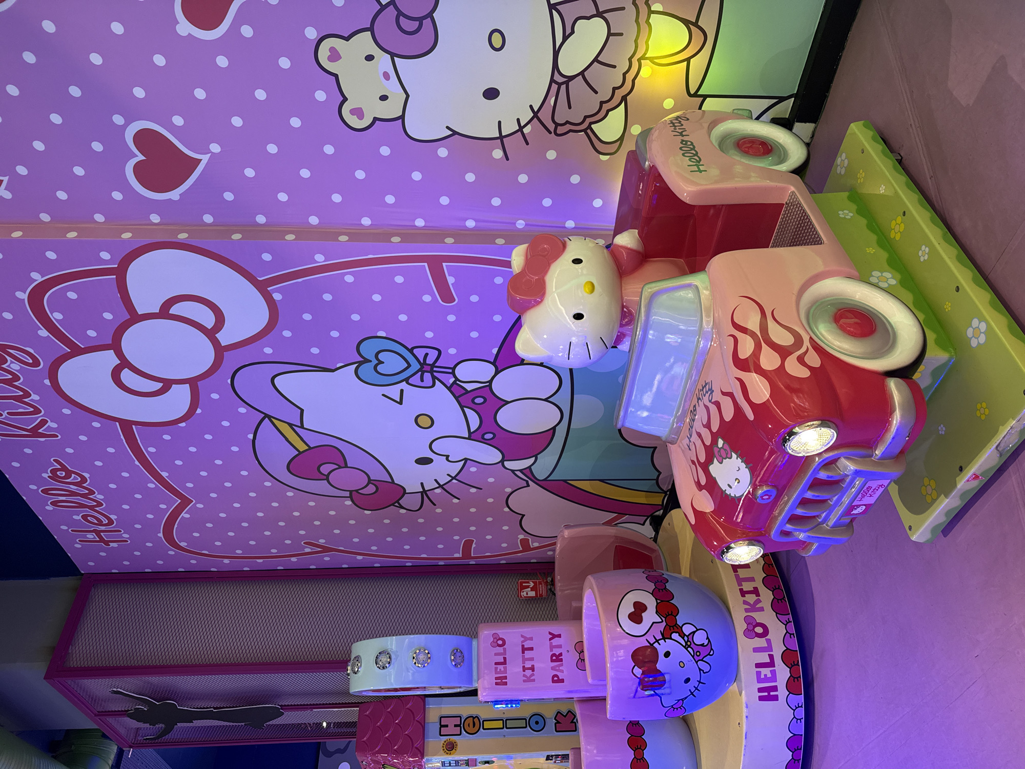 The image captures a brightly lit, pink-themed Hello Kitty-themed area, likely an amusement or entertainment venue. In the foreground, a small, child-sized rideable car, designed to resemble a classic pink and red vehicle with flame decals, is prominently featured. A plastic figure of Hello Kitty sits in the driver's seat of the car.
To the left of the car, a round platform displays several pink cups decorated with Hello Kitty imagery. Behind these cups, a tall, vertical structure with a mesh front and a pink frame stands, featuring a silhouette of a person in a dynamic pose on its side. Another, narrower vertical structure to the left of this one, also pink and mesh, has "HELLO KITTY PARTY" written on it in pink lettering.
The background is dominated by a large mural or backdrop decorated with pink and white polka dots. This backdrop features large illustrations of Hello Kitty, including a depiction of her with a large bow and another, larger image of her wearing a frilly skirt, accompanied by a teddy bear. The overall aesthetic is whimsical and child-oriented, bathed in a soft, purple-pink light.
The image does not contain any explicit information or visual cues that would definitively place it within the city of Üsküdar, Turkey. However, if this is a hypothetical scenario where the location is to be assumed, it would be a commercial entertainment space in Üsküdar.