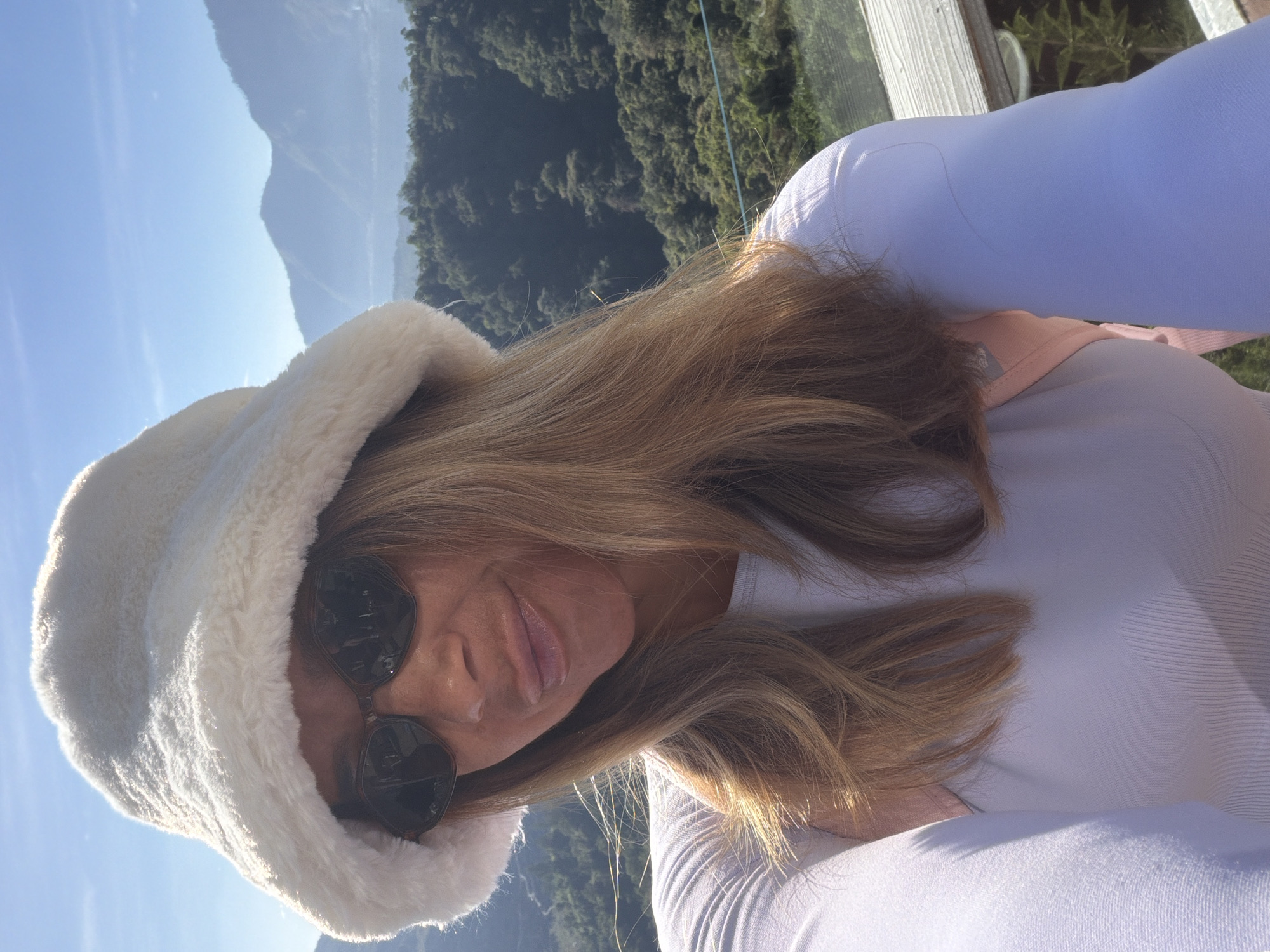 A selfie shows a woman wearing a white fuzzy bucket hat and sunglasses. She is smiling slightly and has blonde hair. She is wearing a light purple long-sleeved shirt. Behind her, a lush green, mountainous landscape is visible under a clear blue sky. The scene is likely in Kintamani, Indonesia, a region known for its scenic highlands and volcanic terrain.