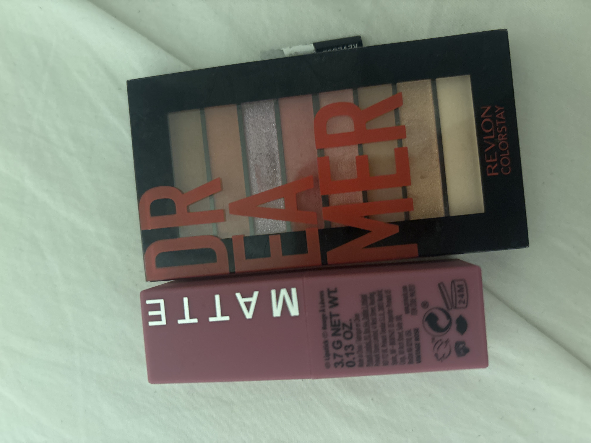 The image shows a top-down view of a pink lipstick and an eyeshadow palette on a white, textured surface. The lipstick tube is upright and positioned to the left, with the word "MATTE" printed vertically in white letters. Below the word "MATTE," there is text detailing the lipstick's weight (3.7 G NET WT. 0.13 OZ.), country of origin, and a symbol indicating a 24-month shelf life. The eyeshadow palette, to the right of the lipstick, is black with the word "DREAMER" printed in large, red, raised letters across the top. Below the word "DREAMER," there are eight eyeshadow pans, varying in shades of pink, coral, and gold, with some pans having a shimmery finish. The bottom of the palette features the Revlon Colorstay logo in red text. The overall scene is well-lit, and the focus is sharp on the products. There is no indication of the location being Dubai, United Arab Emirates.