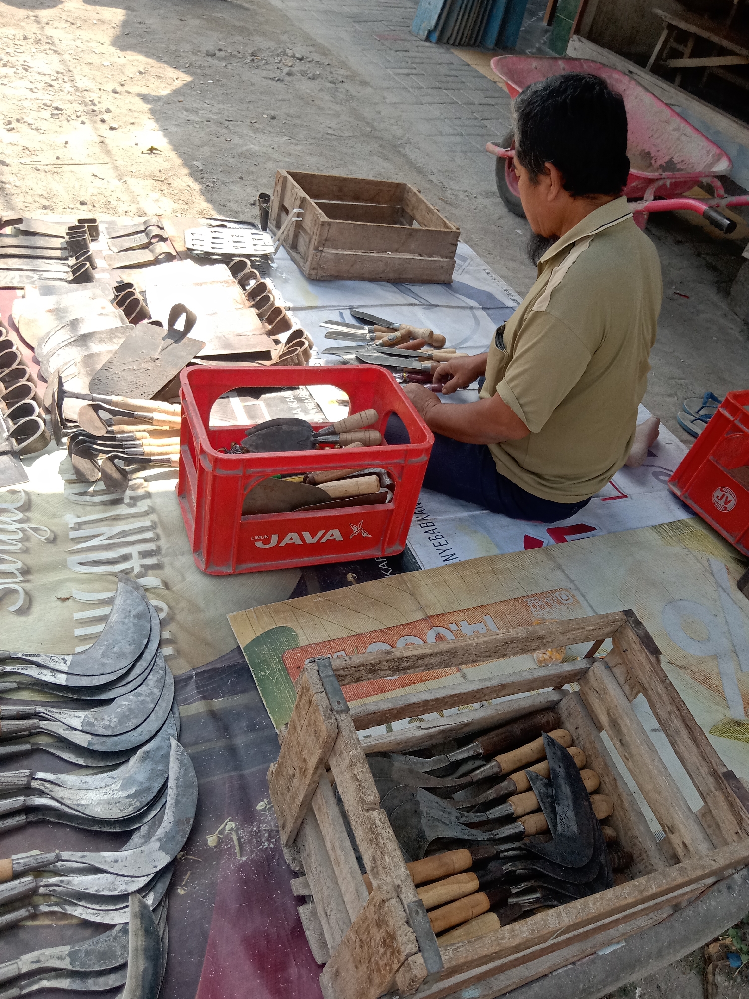 In the city of Klepek, Indonesia, a person is sitting on the ground next to a collection of farming tools. The person, dressed in a tan polo shirt and dark pants, is focused on examining or preparing some of the tools, which are laid out on a mat. These tools include a variety of sickles with curved blades and wooden handles, as well as hoes and other implements. Some of the tools are neatly arranged in a red plastic crate and a wooden crate. The background shows a dusty outdoor area, possibly a market or roadside stall, with a red wheelbarrow visible further in the distance. The overall scene depicts a vendor or craftsman working with agricultural tools in an open-air setting.