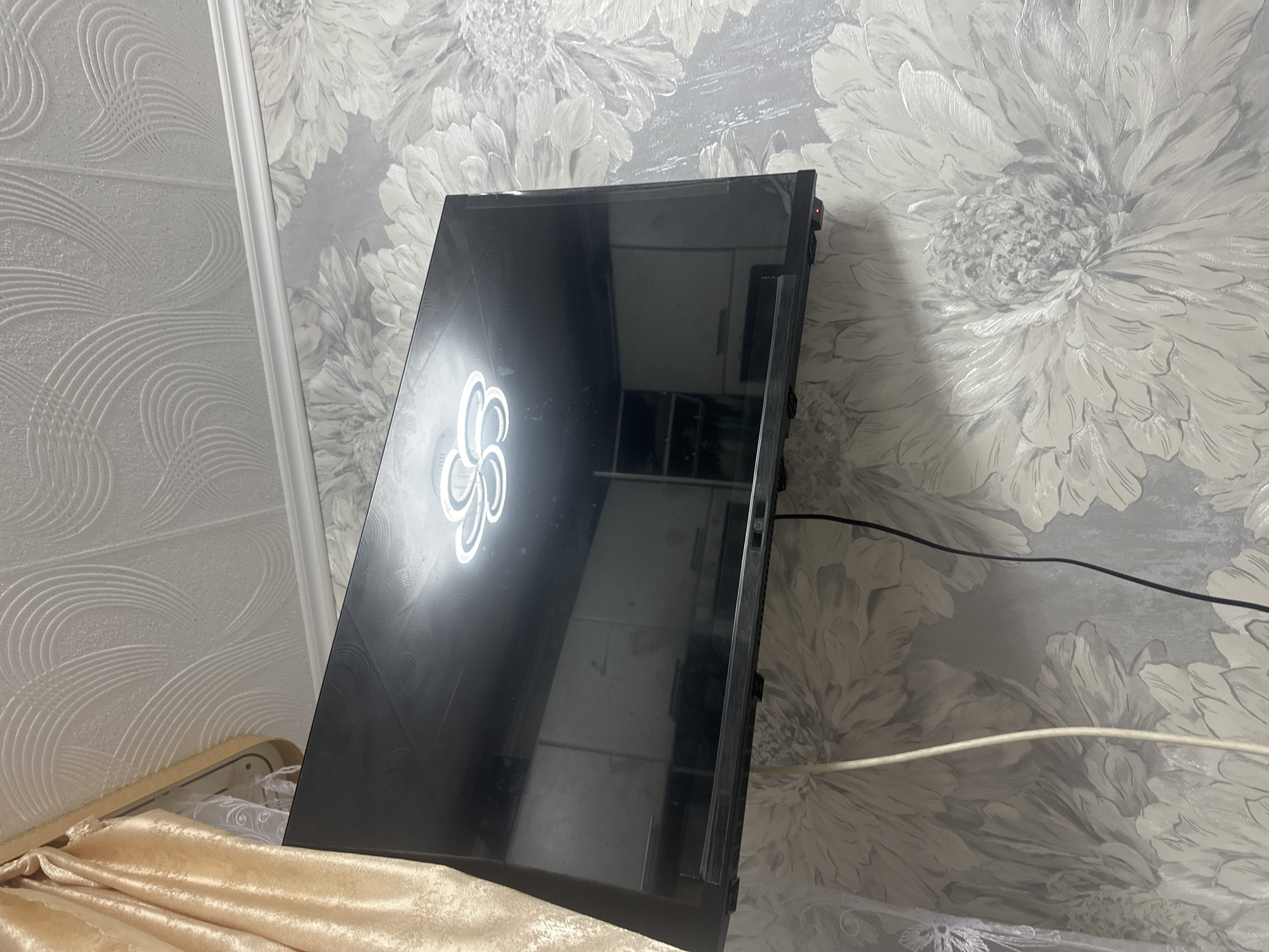 A black flat-screen television is mounted on a wall covered in a floral wallpaper. The television screen reflects a kitchen or cabinet area with shelves. A light fixture or logo is visible on the screen, possibly indicating the TV is on or in standby. To the left of the television, a cream-colored curtain hangs from a rod. White and black cables are visible hanging down the wall from the television. The ceiling above the television has a textured, patterned design. The image appears to be taken indoors, likely in a residential setting.