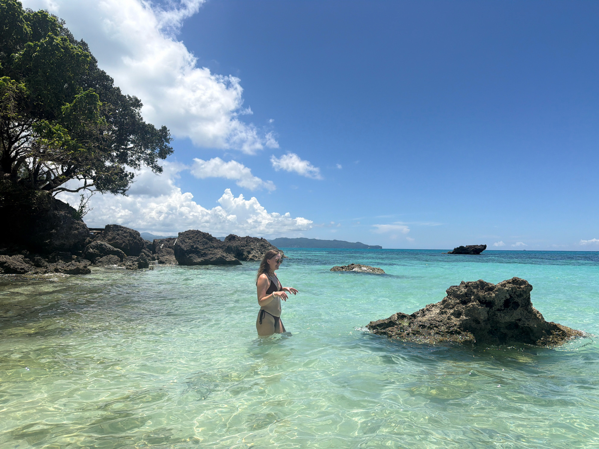 In Malay, Philippines, a woman stands in shallow, crystal-clear turquoise water on a sunny day. She is wearing a brown bikini. To her left, dark, jagged rocks emerge from the water, with a large tree overhanging them, its branches reaching towards the sky. In the background, the turquoise ocean stretches to the horizon, meeting a bright blue sky dotted with fluffy white clouds. Several more rock formations are scattered in the water, and a distant island can be seen on the horizon. The water is so clear that the sandy bottom and ripples on the surface are visible.
