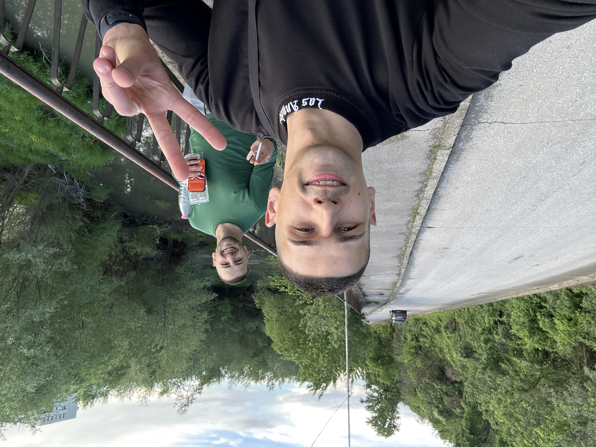 The image shows two men on a bridge. The man closest to the camera is holding up a peace sign. Both men appear to be of similar age and ethnicity. The man further away is holding what appears to be a cigarette and a beverage. The scene is located in Lovech, Bulgaria. There is a river and lush vegetation visible. A gray road and trees are on the right side of the image. A single car is on the road. A building is visible in the distance.