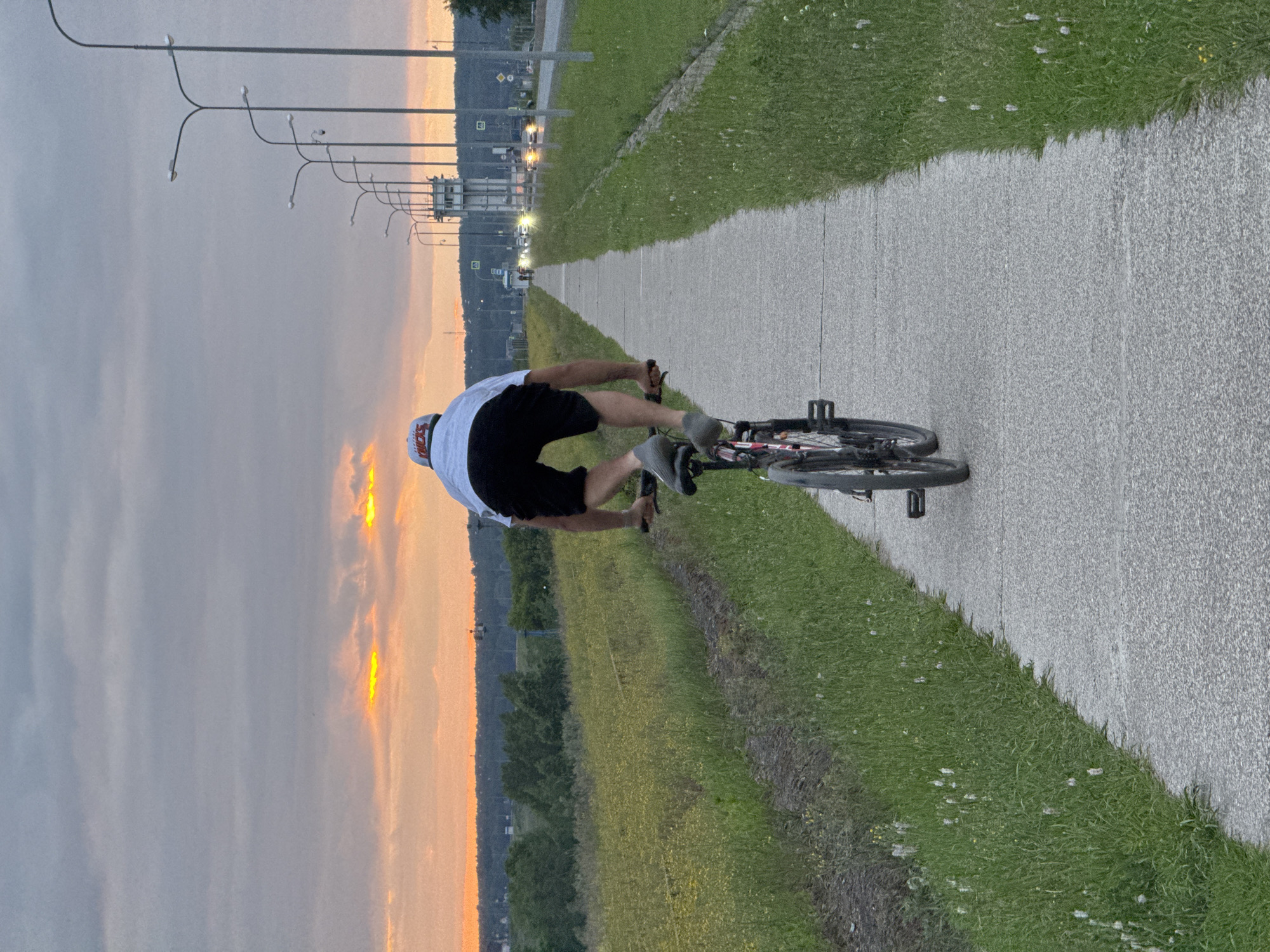 An adult male wearing a white t-shirt and black shorts is riding a bicycle backwards on a paved path. He is wearing a white baseball cap. The path is bordered by a grassy area on the left and a road with streetlights and cars on the right. The sky is cloudy with streaks of orange and yellow from a sunset. In the distance, there is a body of water and some structures. The scene appears to be on the embankment of the Kama River in Chaikovsky, Russia.