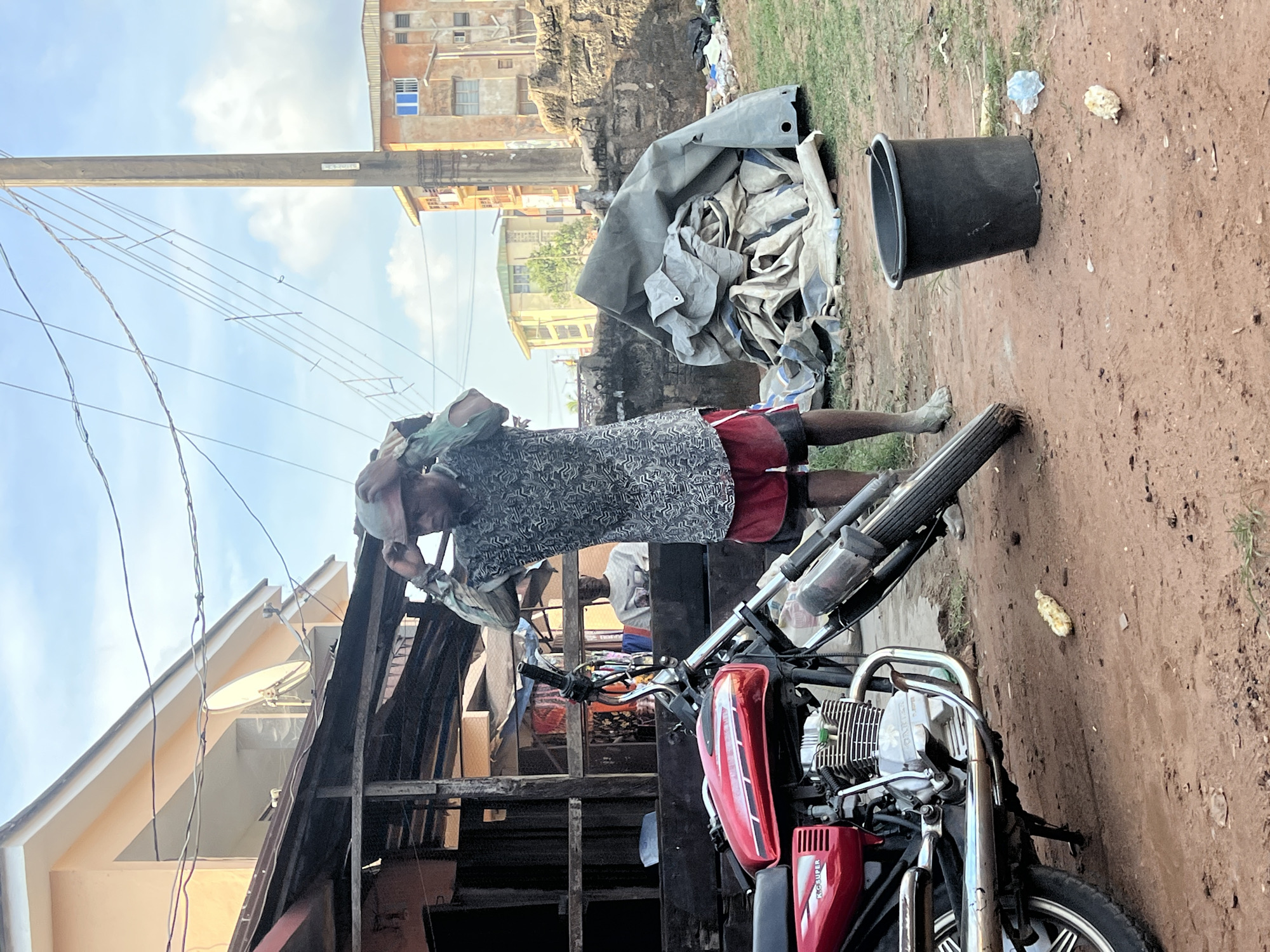 In Nkpor, Nigeria, a man is captured mid-action, adjusting a helmet over his head while standing next to a red motorcycle. He is wearing a patterned shirt and red shorts, with his legs bare except for what appear to be muddy feet or footwear. The motorcycle is parked on a dirt road. In the background, a rustic wooden structure stands, with another person visible inside. Further back, a pile of what appears to be tarpaulins or large fabric sheets is heaped near a low stone wall. The environment suggests an outdoor urban setting, with buildings and utility poles visible under a partly cloudy sky. A black bucket and some scattered debris are also present on the ground.