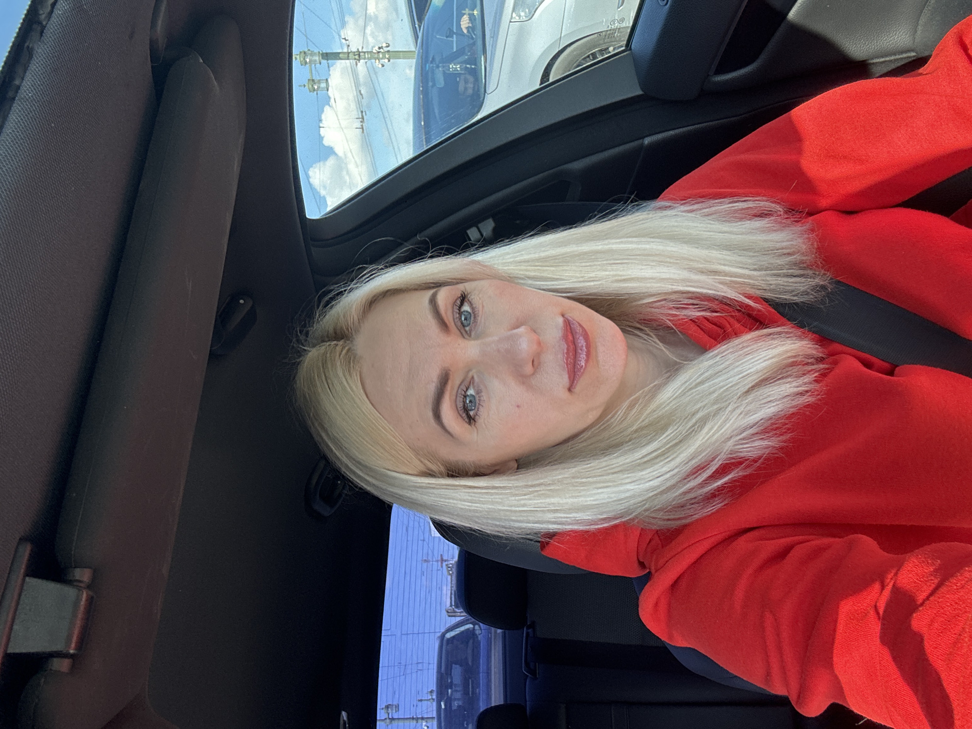 A woman with long, blonde hair is inside a car. She is wearing a red hoodie and a seatbelt. The car is parked in what appears to be a parking lot. Outside the car window, other vehicles and utility poles are visible under a partly cloudy sky. The location in the city is Unknown, Unknown.