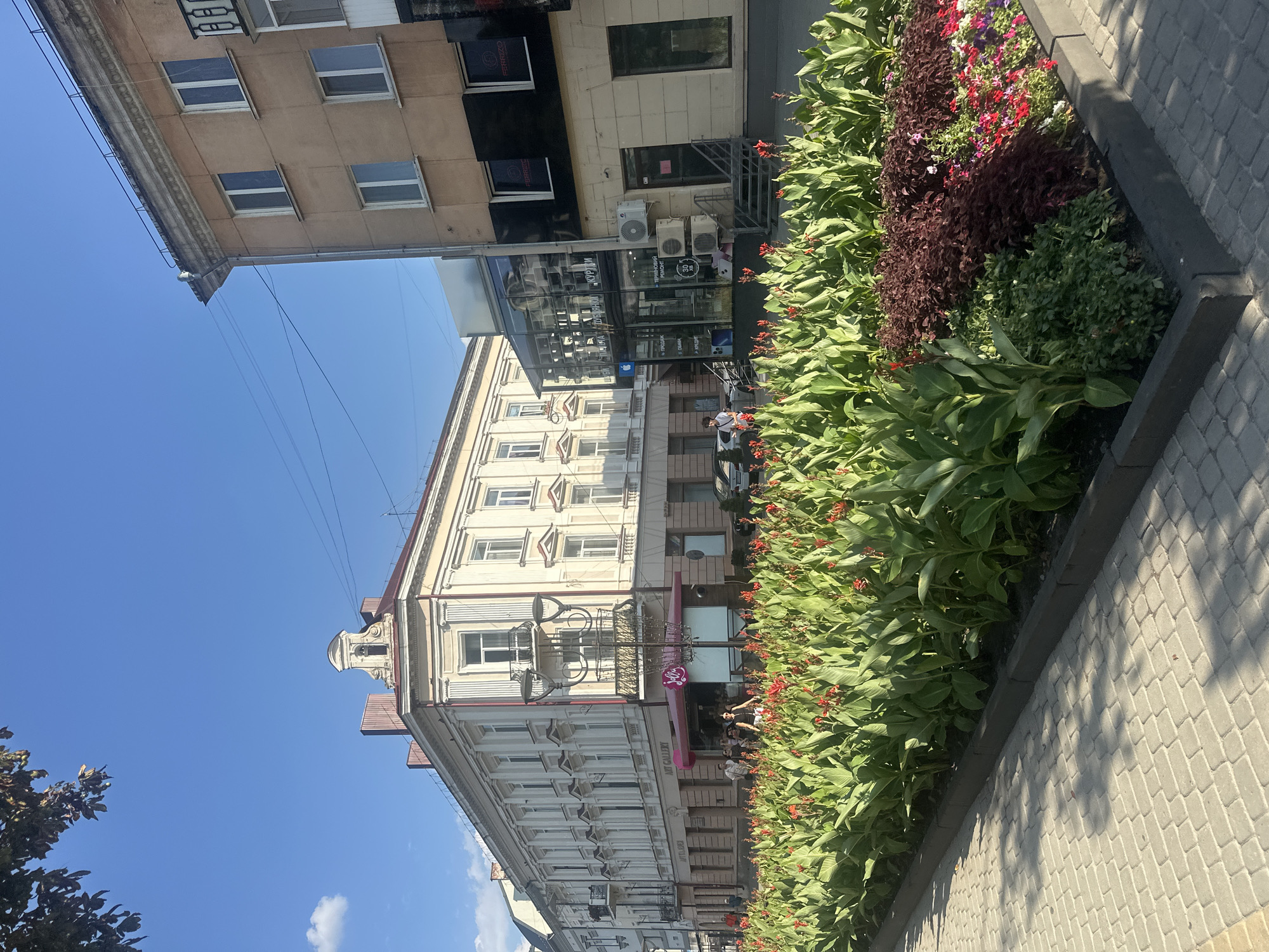 The image captures a vibrant street scene in Ternopil, Ukraine, on a sunny day. In the foreground, a meticulously maintained flower bed bursts with lush green foliage and numerous red and pink blooms, bordered by grey paving stones. Beyond the flowers, a row of buildings lines the street. The building on the left is a light-colored, multi-story structure with ornate architectural details, large windows, and balconies. A pink awning shades a storefront with the signage "Art Gallery" and "Jolli." Several people are visible walking or standing near the entrance to this establishment. To the right, a more modern, beige and black building features large glass windows, air conditioning units, and various signage, including "FERRIZO" visible on the upper windows. A street lamp stands prominently in front of the "Art Gallery." The sky above is a clear blue with a few wispy clouds. The scene suggests a lively urban environment with a focus on public greenery and commercial activity.