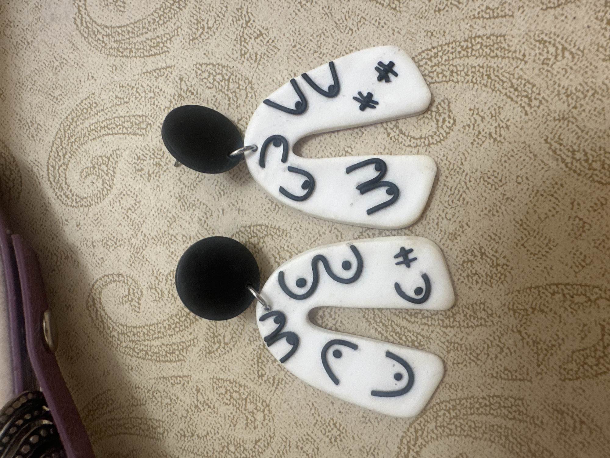 Two handcrafted earrings are displayed against a textured, light brown or beige surface adorned with a subtle, repeating paisley pattern. Each earring comprises a matte black spherical or disc-shaped top, from which a white, inverted U-shaped (arch) component is suspended by a small silver-colored jump ring.
The white arch components are made of polymer clay or a similar craft material and feature various abstract designs rendered in black.
The left earring exhibits multiple stylized "U" shapes with central dots, resembling breasts. Specific designs include: two stacked "U" shapes with dots on the upper left arm, a single "U" shape with a dot on the lower left arm, a wavy line with two dots on the upper right arm, and a "C" shape with a dot next to a hash symbol on the lower right arm.
The right earring also features similar stylized designs: two stacked "U" shapes with dots on the upper left arm, a "W" shape with three dots on the lower left arm, two stacked "V" shapes with dots on the upper right arm, and two asterisk-like symbols on the lower right arm.
In the upper left portion of the image, a segment of a dark, textured object, possibly metallic jewelry, is visible. Adjacent to and partially covering this object is a strip of purple material, which includes a small, circular, silver-colored snap fastener. The overall lighting is even, suggesting an indoor setting.