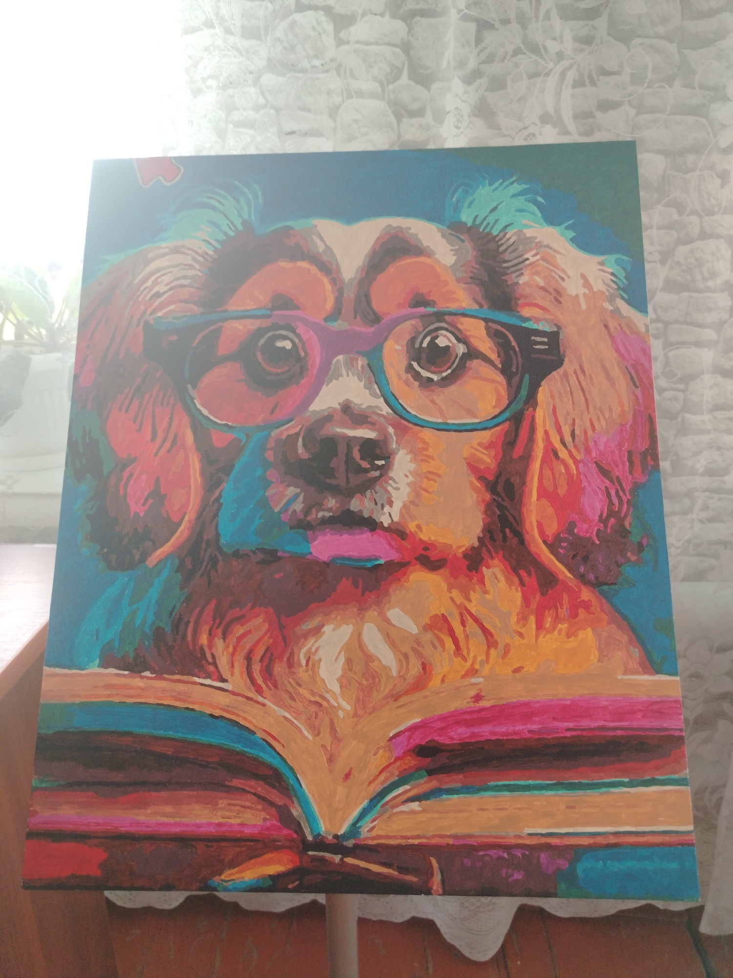 The image is a close-up, top-down view of a painting depicting a dog wearing glasses and reading a book. The painting uses vibrant, saturated colors with a distinct, blocky brushstroke style, reminiscent of paint-by-numbers art. The dog's fur is rendered in oranges, reds, and blues, with green accents on its head. It wears purple-rimmed glasses and has its tongue slightly sticking out. The open book beneath the dog is also vividly colored with stripes of pink, orange, blue, and red. The painting is displayed on a surface that appears to be a table covered with a white lace tablecloth. The background shows a textured, light-colored wall. The image is partially obscured by a strong light source, likely a window, which creates a bright glare in the upper left corner and washes out some details of the painting. There are no people visible in the image. The painting itself is the sole subject. The provided information about the location in Klyuchi, Russia, does not correspond to any visible elements within the image itself, which solely depicts the painting and its immediate surroundings.