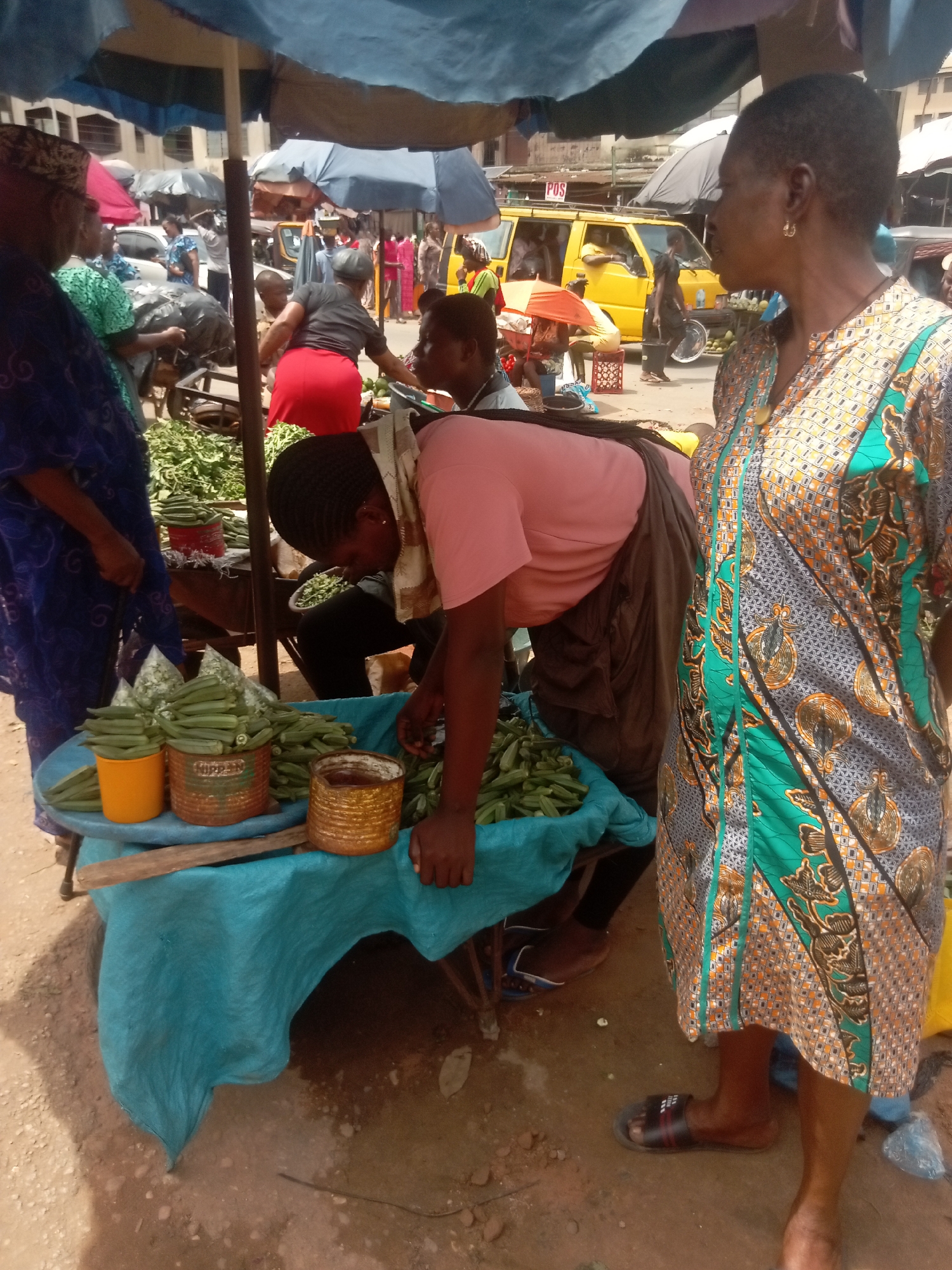 On a street in Onitsha, Nigeria, a marketplace scene unfolds under the shade of umbrellas. A woman in a patterned green and gray dress stands observing. Another woman in a pink shirt sorts okra on a table covered with a blue cloth. Containers of okra are arranged on the table. A third woman in a blue patterned dress stands nearby. Other people, umbrellas, carts, and a yellow vehicle marked "POS" are visible in the background.