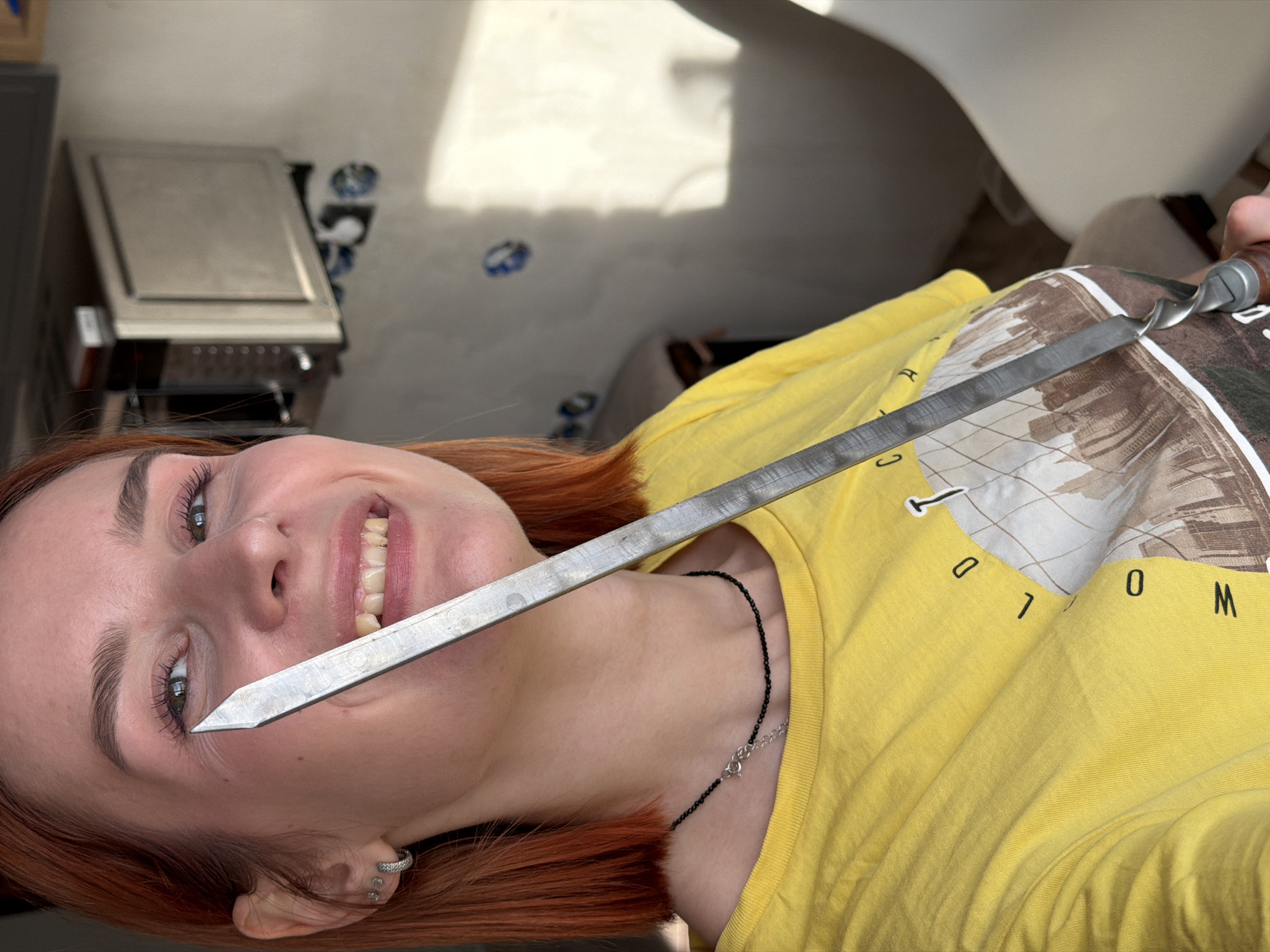 In an indoor setting in Chaikovsky, Russia, a female subject with reddish-orange, shoulder-length hair and light complexion is depicted smiling directly towards the camera. She wears a bright yellow short-sleeved t-shirt featuring a partial graphic design with "OLD TOWN" text, a thin black beaded necklace, and two small silver hoop earrings on her left ear. Her mouth is open in a smile, revealing her teeth. She holds a long, flat, metallic skewer, approximately 40-50 cm in length, with a twisted central section and a dark-colored handle, in her right hand. The pointed tip of the skewer is positioned vertically near her left eye. The background consists of a light-colored, possibly off-white or beige, textured wall. Multiple blue markings are visible on the wall, indicative of electrical outlet or fixture preparations. To the upper right of the subject, a stainless steel appliance, consistent with a microwave or compact oven, is mounted on the wall. Strong natural light illuminates the scene from the right, casting distinct shadows on the background wall.