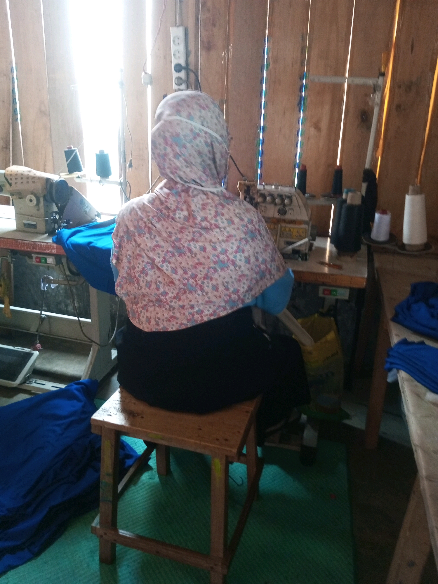 A person wearing a headscarf with a floral pattern and a dark-colored bottom sits on a wooden stool in front of a sewing machine, appearing to be in a workshop. The person's back is to the camera, and they are engaged with the sewing machine. Various spools of thread, one white and several black, are visible on the machine and a nearby shelf. A large piece of blue fabric is draped over the sewing machine table and also lies on the floor. The workshop appears to be constructed with wooden walls and beams. Natural light streams in from an unseen source, creating bright highlights and shadows. The overall impression is of a utilitarian workspace focused on textile production. This scene is located in the city of Kalibeber, Indonesia.