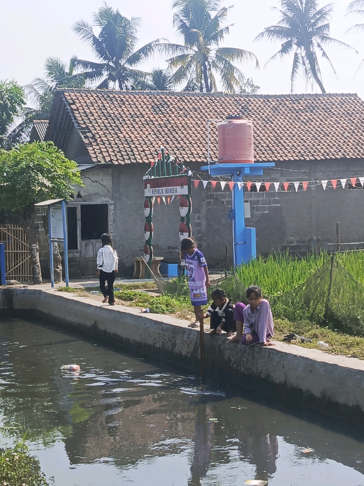 In Rawa Boni, Indonesia, a concrete canal runs through a village. Children sit on the edge of the canal. One child, wearing a purple and blue jersey, holds a stick. Another child, in a light purple shirt, sits next to them, appearing to engage with the stick. A third child, in a black and white shirt, kneels nearby. The canal water is murky, with some floating debris, including a white and red plastic bottle. In the background, a building with a tiled roof stands behind a gate decorated with red and white triangular flags and circular emblems. A large orange water tank is mounted on a blue structure in front of the building. Palm trees and other foliage are visible, suggesting a tropical or subtropical environment. A young woman in a white shirt and dark pants walks away from the canal on a paved path. The overall scene depicts a residential area with a canal, likely a part of the local infrastructure.
