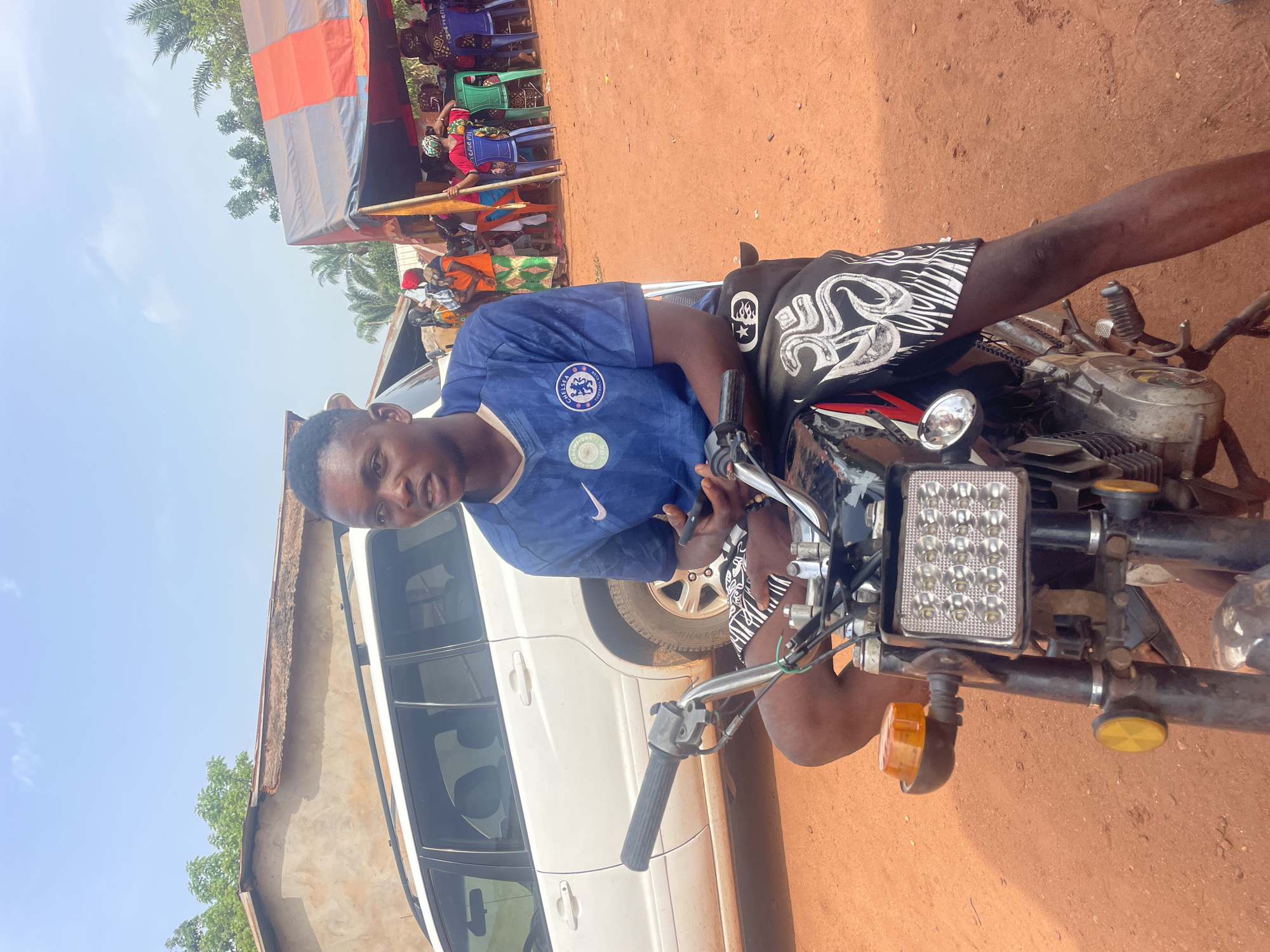 A young man is posing on a motorcycle in Ogbe, Nigeria. He is wearing a blue Chelsea soccer jersey and black shorts with white patterns. The motorcycle is black and chrome, with an aftermarket LED headlight. Behind the motorcycle, there is a white vehicle and a building with a light-colored wall. In the background, people are gathered under a canopy. The ground is red dirt.