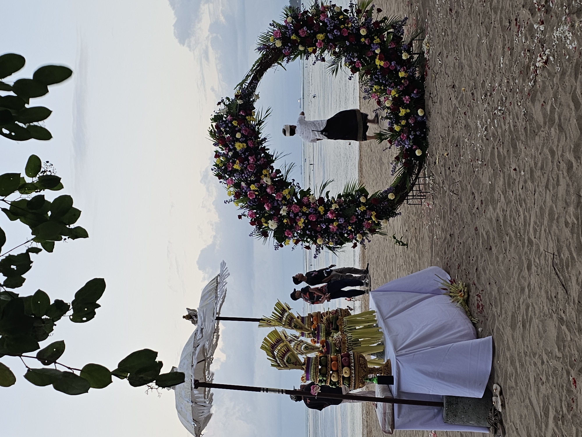 This image depicts a beach scene in Denpasar, Indonesia, likely during a pre-ceremony or setup for an event. In the foreground, a table draped with a white cloth displays offerings, including intricately arranged fruits and what appear to be woven leaf decorations, under two white ceremonial umbrellas. Several people are gathered near the table, observing the setup. To the right, a large circular archway, elaborately decorated with vibrant flowers in shades of pink, yellow, and purple, stands prominently on the sand. A person dressed in traditional Balinese attire, a white shirt and a dark sarong, stands near the arch, facing the ocean and gesturing. The background shows a calm sea with a faint horizon, and the sky is overcast with soft clouds. The beach itself is sandy, with scattered flower petals on the ground, suggesting recent or ongoing preparations. The overall atmosphere is serene and anticipatory.