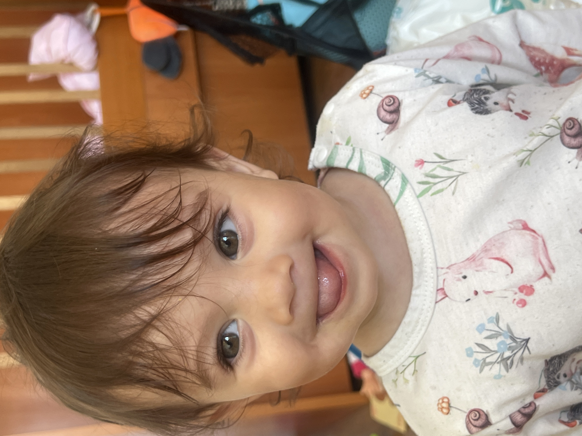 A close-up portrait, taken at an angle, captures a joyful baby looking directly at the camera. The baby has expressive, large dark brown eyes and soft, wispy dark brown hair that frames their face. A cheerful expression is visible, with the baby's mouth slightly open and tongue peeking out, conveying a sense of playfulness and happiness.
The baby is wearing a light-colored top or onesie adorned with a delightful pattern of nature-themed illustrations. These include whimsical pink rabbits, small grey hedgehogs, and brown snails, interspersed with delicate blue and pink flowers and green foliage. The fabric appears soft and comfortable.
The background is softly blurred, revealing warm-toned wooden elements, possibly part of a crib or furniture. Hints of other out-of-focus objects, including an orange item and a black mesh-like material, suggest an indoor setting, likely a nursery or home environment. The lighting appears natural and bright. No specific time of day or weather conditions are discernible, nor are there any visible texts or specific location cues to confirm the Elena, Bulgaria context beyond the prompt's information. The overall mood of the image is one of innocence, warmth, and infantile cheerfulness.