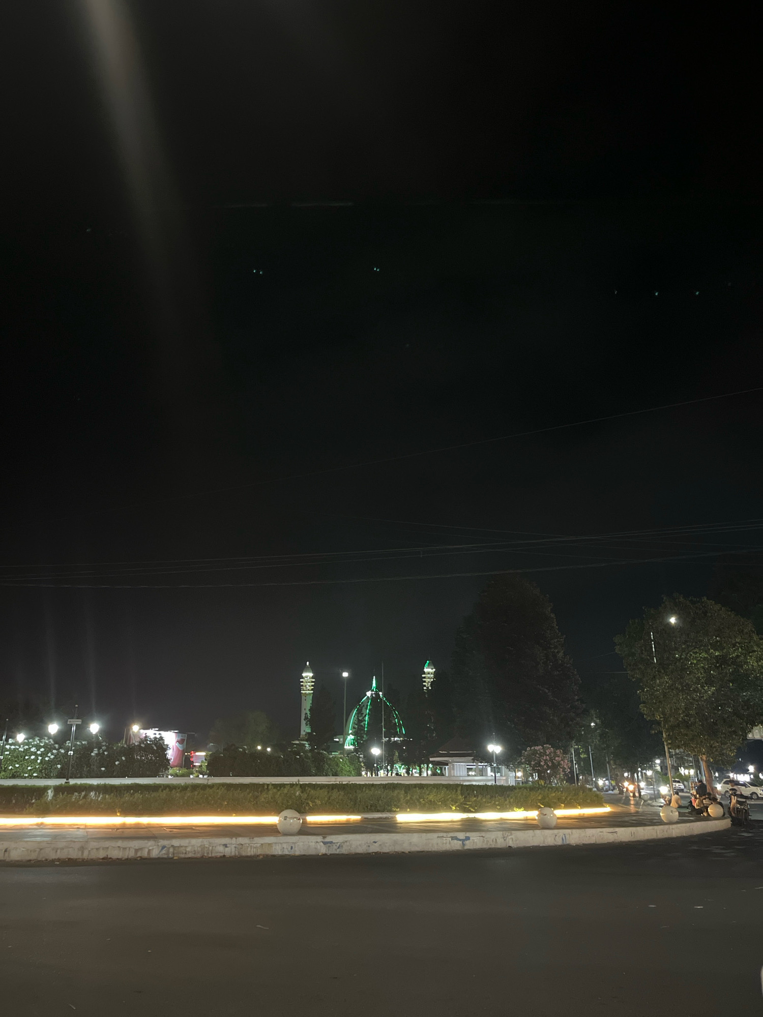 A night-time, horizontal, eye-level, medium shot captures a brightly lit roundabout with long-exposure light trails from passing vehicles in Lumajang, Indonesia. In the background, illuminated buildings and trees are visible, including a prominent green domed structure and a tall, white minaret. The scene is characterized by a dark sky with some atmospheric haze and a single bright beam of light extending from the upper left corner downwards. The foreground shows a dark asphalt road and the edge of the roundabout, which is decorated with white planters. A few motorcycles with riders are parked on the right side of the image, and distant streetlights create a scattered illumination.