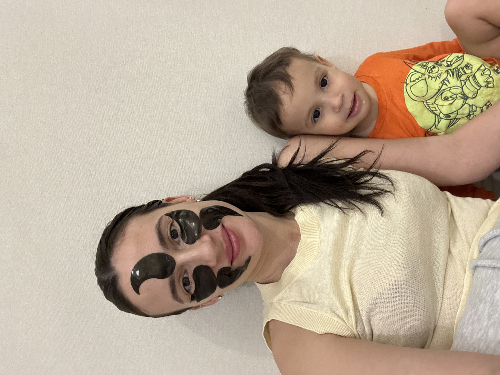 A woman and a child are lying next to each other on a light-colored surface. The woman has dark hair pulled back in a ponytail, and she is wearing black eye patches. She is wearing a light yellow t-shirt. The child has short brown hair and is wearing an orange long-sleeve shirt with a cartoon design. The image appears to be taken indoors against a plain, off-white wall. The location is unknown as it is not possible to determine this from the image.