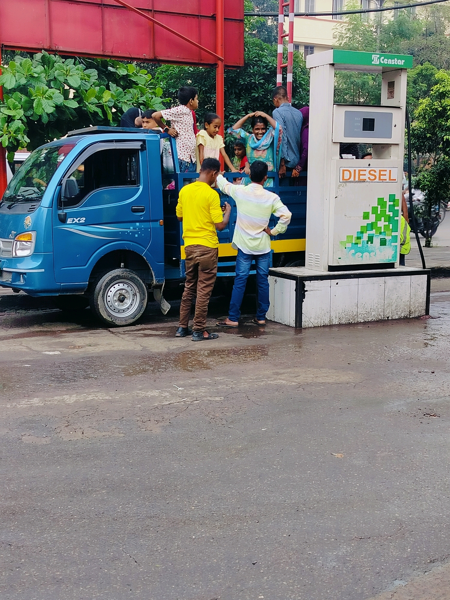 At a roadside gas station in Dhaka, Bangladesh, a blue truck, identified as "EX2", is parked next to a "Censtar" brand diesel pump. Several people, including children, are gathered around and inside the truck. Two men stand on the ground near the truck and the pump. One man, wearing a yellow t-shirt and brown pants, is facing away from the camera, while another man in a striped shirt is seen from the back. The ground is wet, suggesting recent rain. The overall scene depicts a common sight at a fuel station, possibly during busy hours or a period of informal transport.