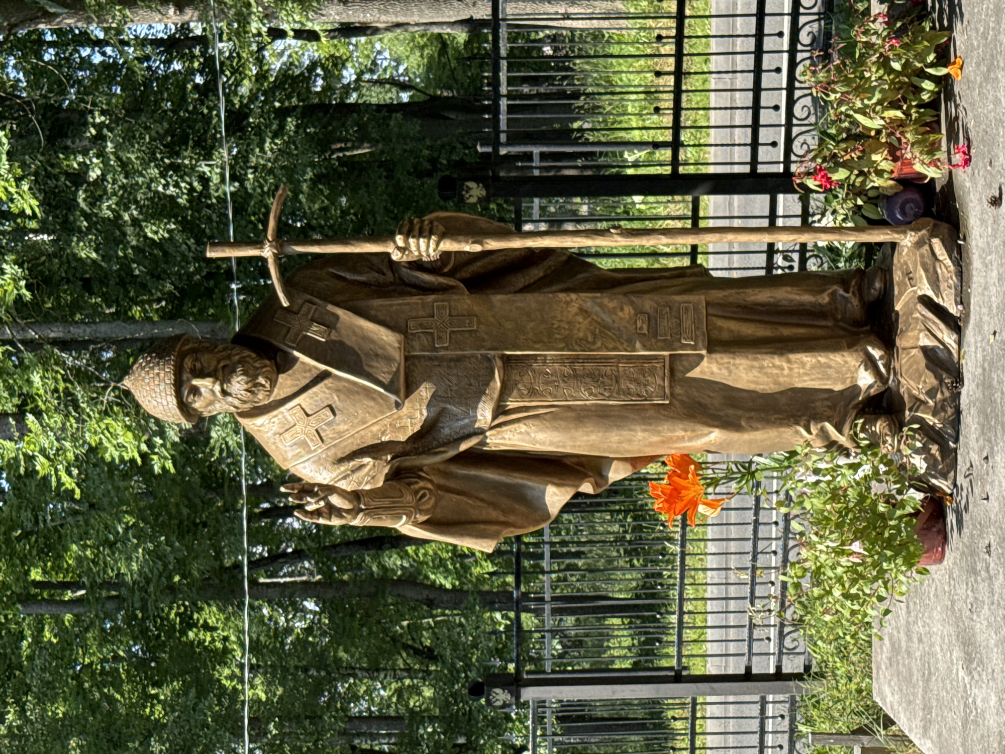 A bronze statue of Saint Nicholas of Myra stands in a cemetery, his gaze directed slightly downwards and to the left. He is depicted wearing a bishop's miter and vestments adorned with crosses and ornate patterns. In his right hand, he holds a crozier, while his left hand is extended outwards. The statue is positioned in front of a black metal fence, with a paved walkway and lush green trees visible beyond it. Potted plants with red and orange flowers are placed near the base of the statue, adding a touch of color to the scene. The lighting suggests it is daytime, with the sun casting shadows and highlighting the bronze surface. The overall atmosphere is solemn and reverent, fitting for a memorial. The statue is likely located in Lomonosov, Russia, as indicated by the context.