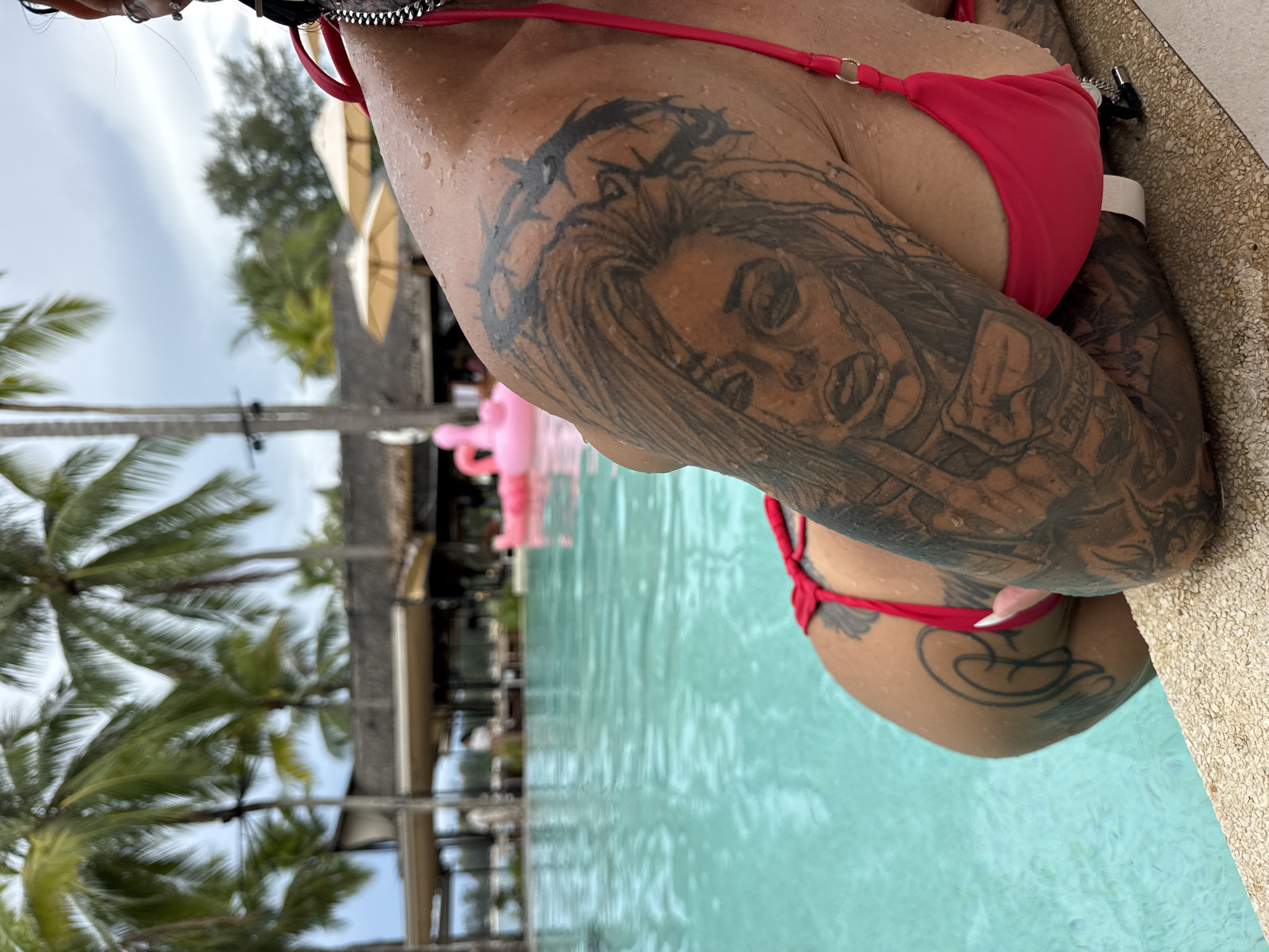 In Choeng Thale, Thailand, a person is partially submerged in a swimming pool, with their upper body and tattoos prominently displayed. The person is wearing a pink bikini. Their left arm, adorned with extensive black ink tattoos, rests on the edge of the pool. Visible tattoos include a detailed portrait of a face wearing a crown of thorns on the upper arm and shoulder, and other designs on the forearm and part of the back. Water droplets are visible on the person's skin. The background reveals a tropical setting with palm trees, a building with a thatched roof, umbrellas, and a pink inflatable flamingo in the pool. The water in the pool is a clear turquoise color.