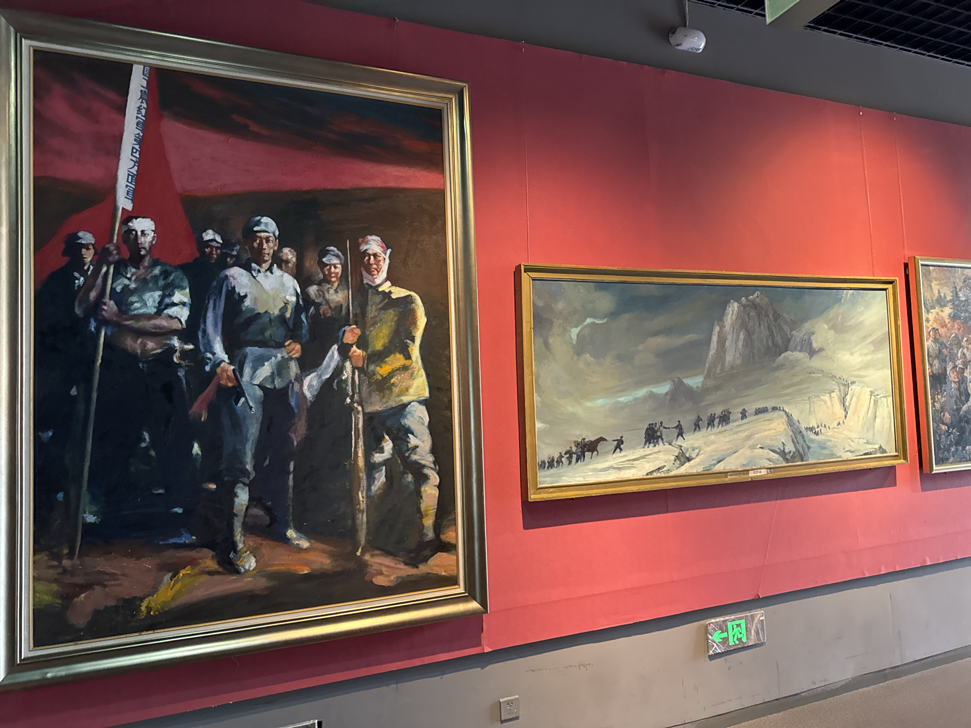 The image shows a display of paintings on a red wall in what appears to be a museum or gallery.
The largest painting, framed in silver and gold, depicts a group of soldiers, likely from a historical army. They are carrying rifles and a flag with writing on it. The background of the painting is primarily red.
To the right of this painting are two smaller, rectangular paintings with gold frames. One depicts a landscape with figures walking along a path towards a mountain. The third painting shows people in action within a detailed scene.
The wall is painted a bright, matte red, and below the paintings, the wall transitions to a grey color. At the bottom right of the wall is a green emergency exit sign.
Based on the limited information, the location is likely within the West Station Subdistrict in China, given the nature of the artwork.