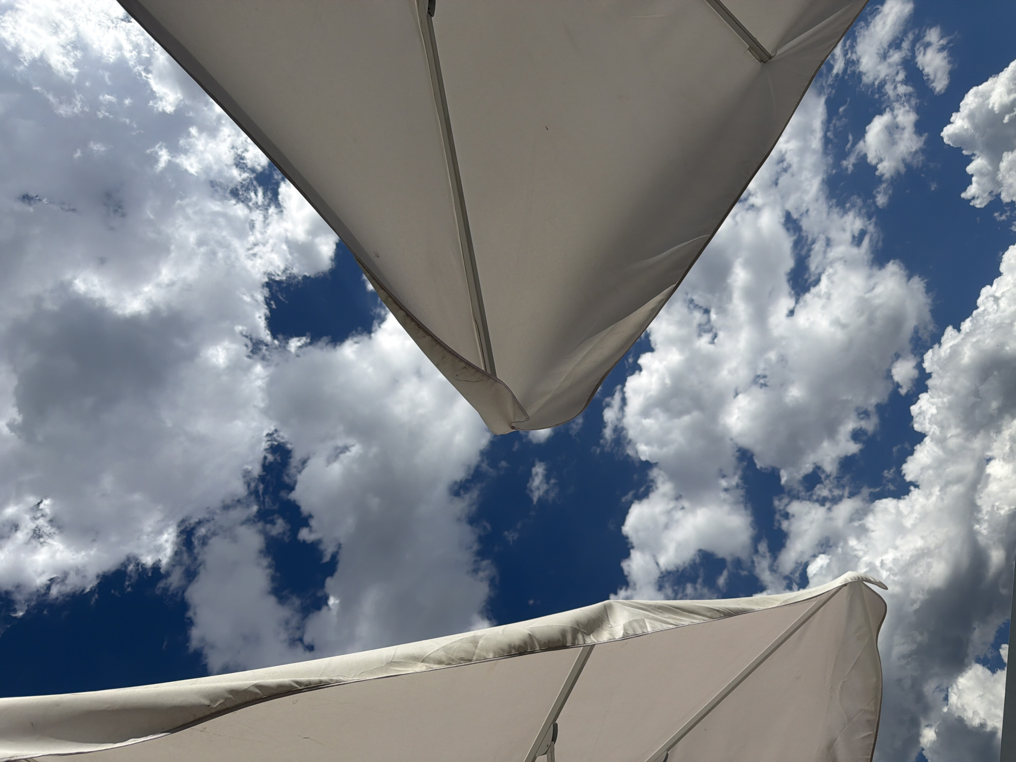 The image shows a close-up, low-angle view of two white patio umbrellas against a bright blue sky filled with fluffy white clouds. The umbrellas are angled upwards, with their fabric taut and supported by unseen structures. The left umbrella is larger and closer to the viewer, showing the curved edge and a seam. The right umbrella is smaller and further away, appearing as a sharp diagonal line against the sky. The scene is devoid of people. The overall impression is one of an outdoor setting, possibly a cafe or a park, on a clear, cloudy day. The specific location in Sofia, Bulgaria is not discernible from the visual elements of the image.
