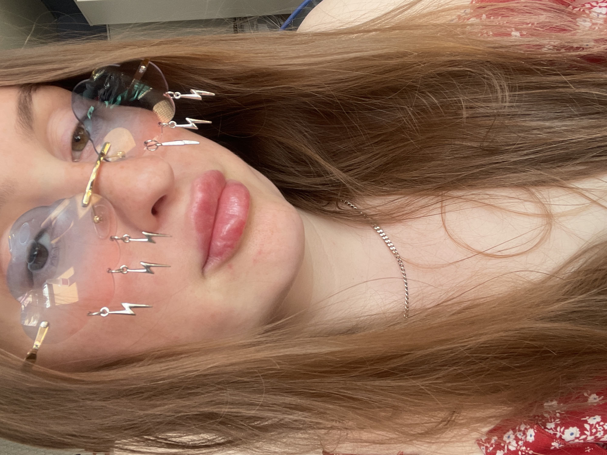 A close-up, chest-up shot shows a person wearing cloud-shaped, rimless sunglasses with several lightning bolt charms dangling from them. The sunglasses have a blue and green iridescent tint. The person has light skin with some redness on their cheeks and nose, and their lips are full and glossy. They have long, straight, reddish-brown hair that falls around their face and shoulders. Around their neck, they wear a thin, silver chain necklace. They are wearing a red top with a white floral pattern. The background is out of focus and appears to be indoors, with a white wall and a glimpse of what might be furniture. This image was captured in Сырково, Russia.