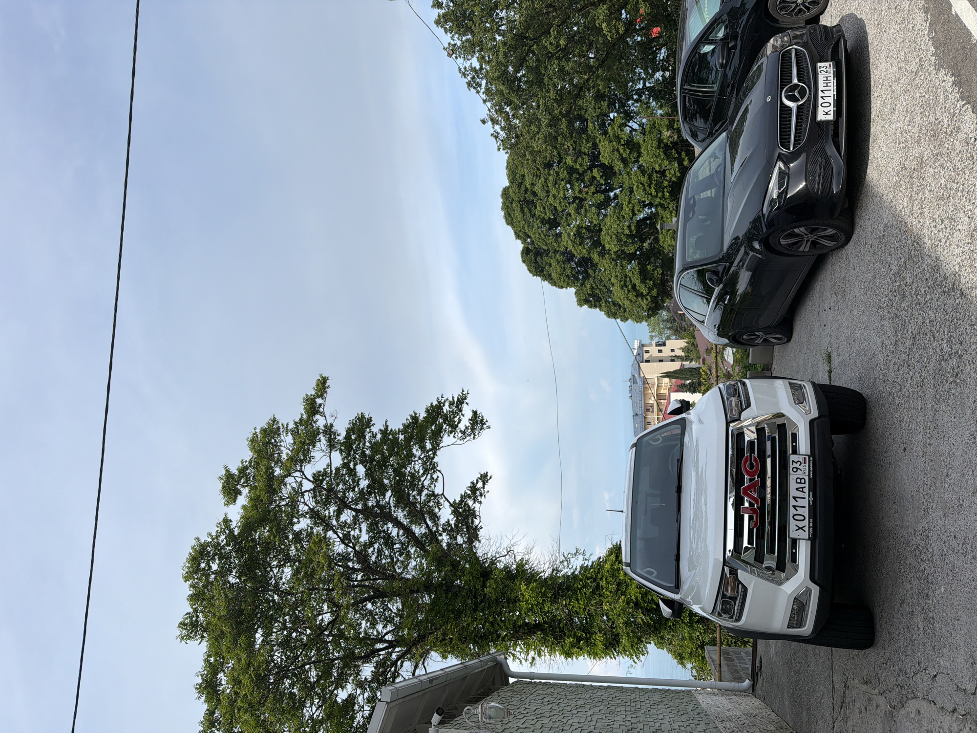 A white JAC JS6 SUV is parked in the foreground on a paved surface, angled towards the viewer. The SUV has a chrome grille with a red JAC logo and a license plate reading "X 011 AB 93". To the right of the SUV, a black Mercedes-Benz C-Class sedan is parked, also facing forward, with its license plate reading "K 011 HH 23". Behind the Mercedes, a portion of another black car is visible. A large green tree with dense foliage dominates the left side of the frame, with its trunk covered in ivy. In the background, buildings and the sea are visible. The sky is partly cloudy. The scene appears to be in Sochi, Russia, given the license plate format and the architecture visible.