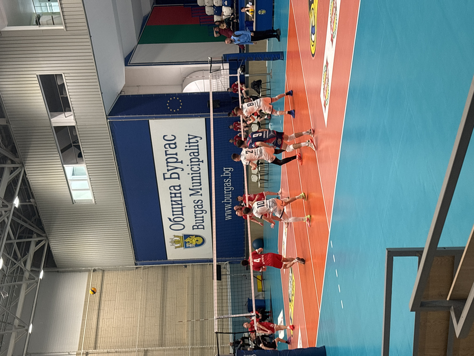 The image shows a volleyball game in progress at a sporting venue, likely located in Burgas, Bulgaria, given the "Община Бургас Burgas Municipality" banner in the background. The volleyball net divides the court into two sides.
One team, wearing predominantly white jerseys with dark accents, is positioned near the center of the court and in the middle of a play. Their opponents, wearing red jerseys, are on the opposite side of the net. A volleyball is in the air near the net. Players on both sides are positioned as though mid-play, indicating active engagement in the game.
Spectators are visible in the background. The lighting appears to be artificial, as is usual in an indoor sports arena. The walls and ceiling show typical design elements for such a venue. The floor of the court is colored, with orange and blue sections visible.