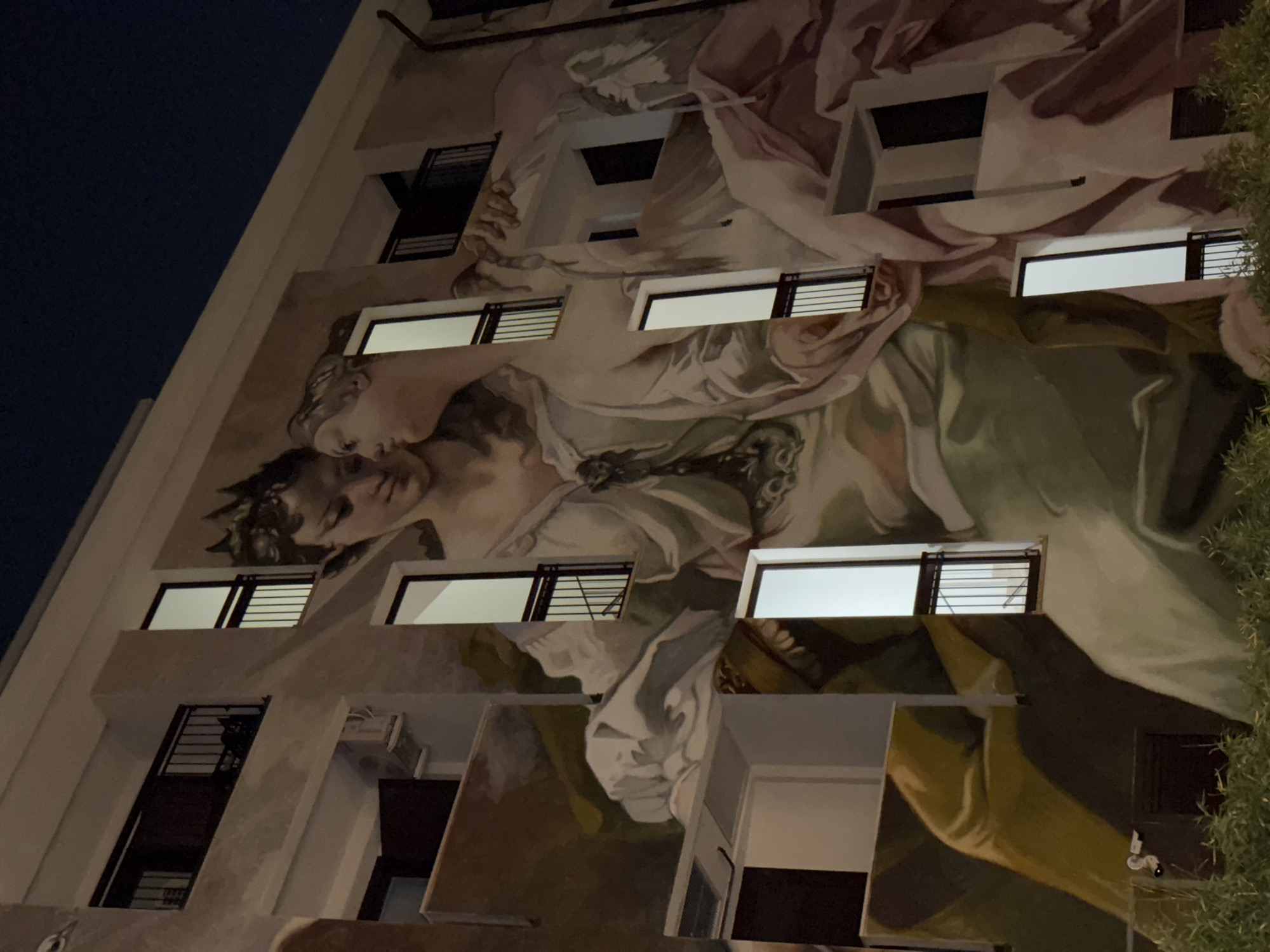 A low-angle shot shows the facade of a building at night in Milan, Italy. The building has a mural depicting what appears to be two figures in a classical or Renaissance style embracing. Windows are interspersed throughout the mural, some with blinds and lights visible. The sky is dark blue or black in the upper-left corner. Green foliage is visible along the right edge of the frame.